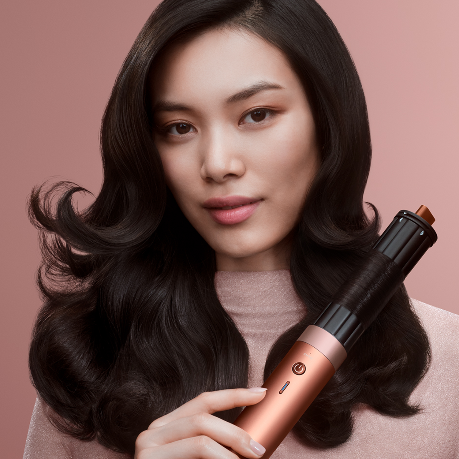A woman with dark hair holds a hair curler. She wears a pink top, and the background is a soft pink.