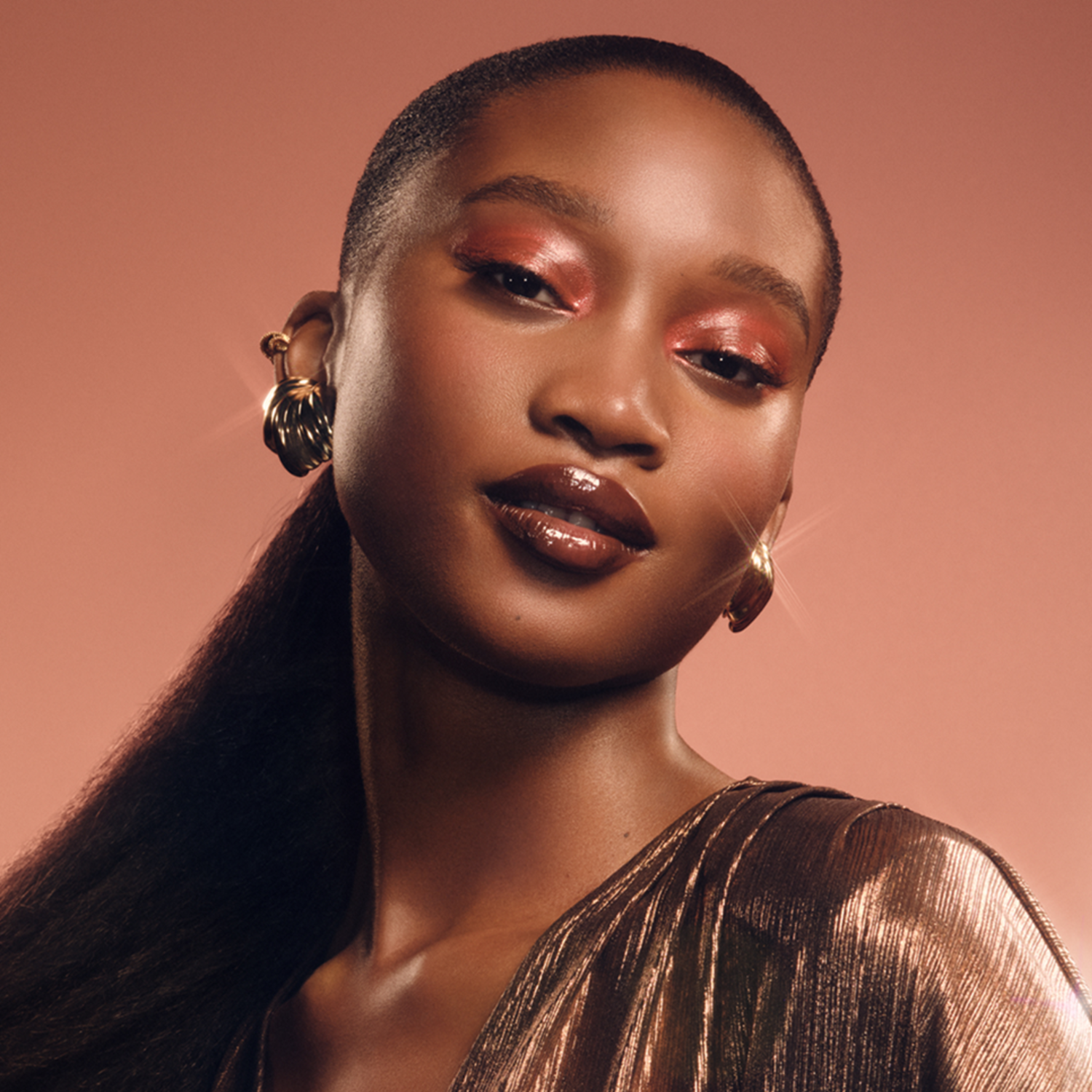 A woman with dark skin and glamorous makeup, gold earrings, and a gold dress, poses against a peach background.
