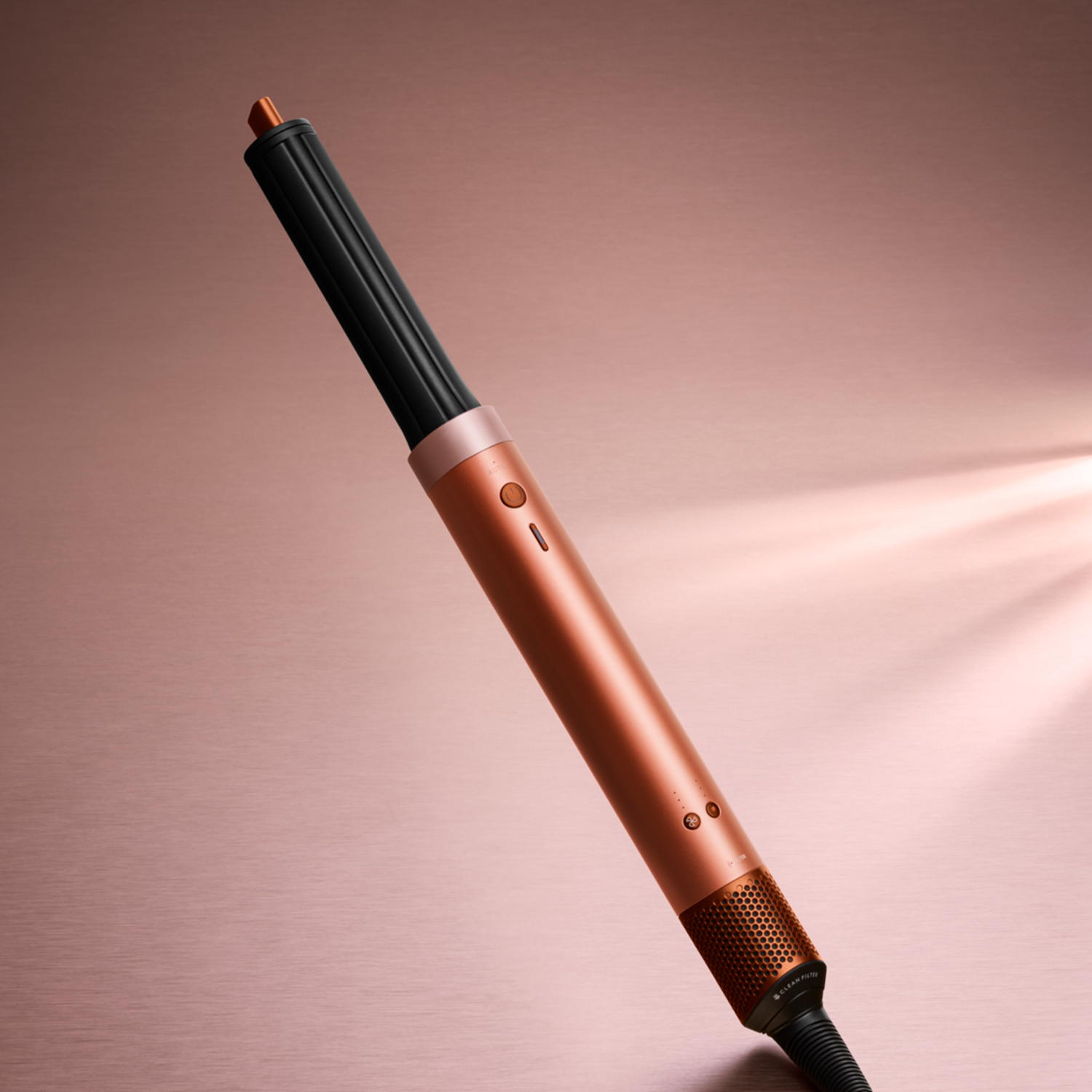 A rose gold hair styling tool with a black cylindrical part on top. It has buttons and a mesh at the bottom on a pink background.