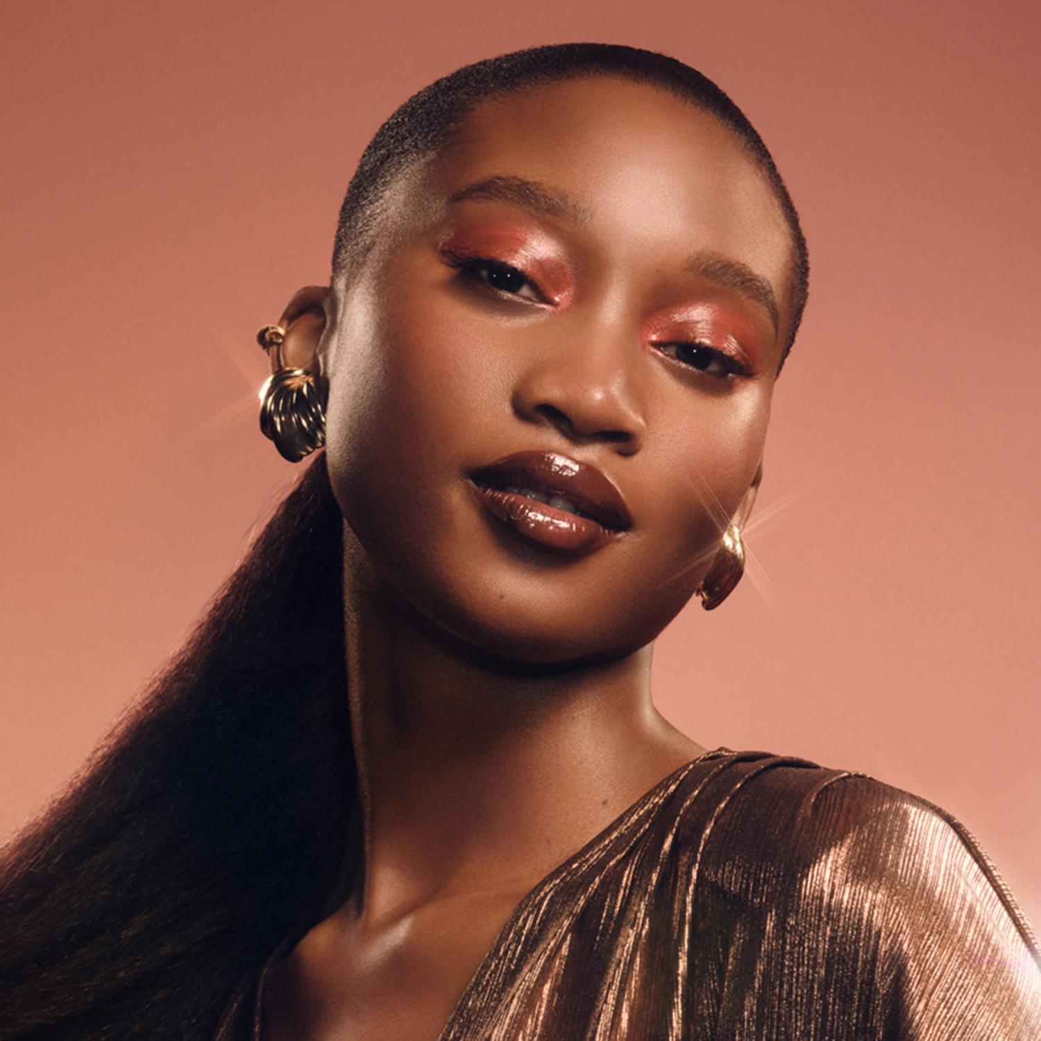 A woman with dark skin, wearing gold earrings and a dress. She has red eye shadow and glossy lips. The background is pink.