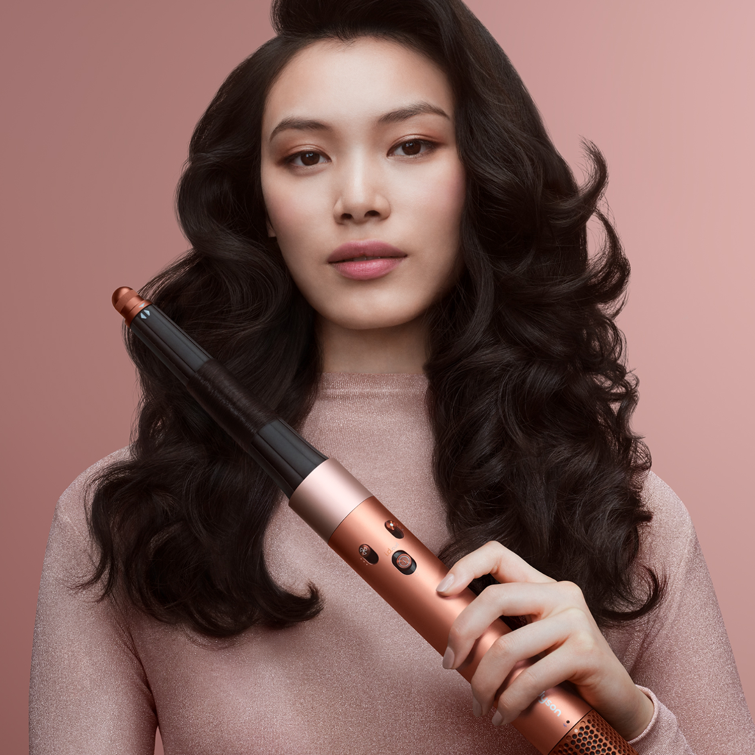 A woman with dark hair holds a copper-colored hair styling tool. She wears a pink top, set against a pink background.