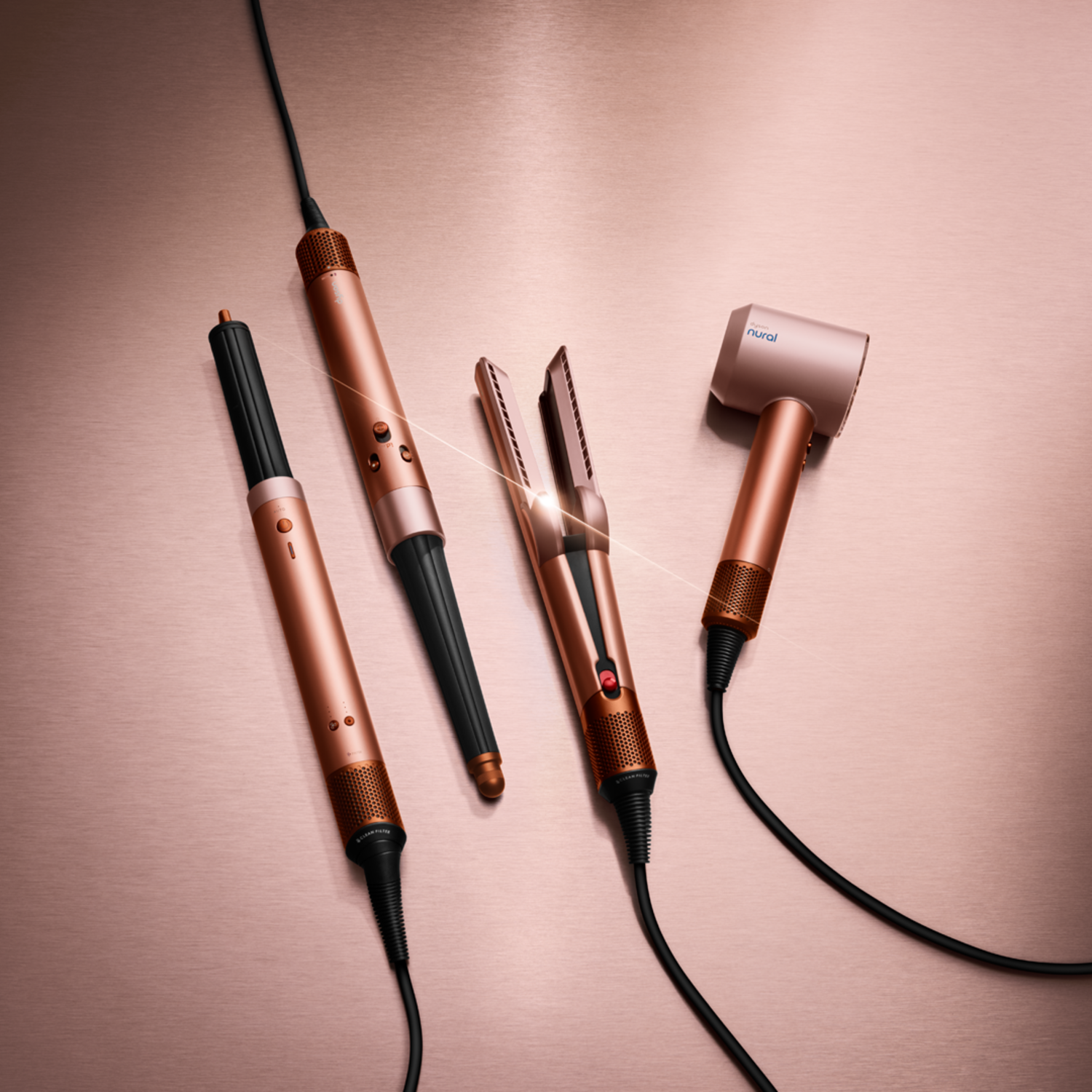 Rose gold hair styling tools on a rose gold surface. Includes a curling wand, hair straightener, and hair dryer with black cables.