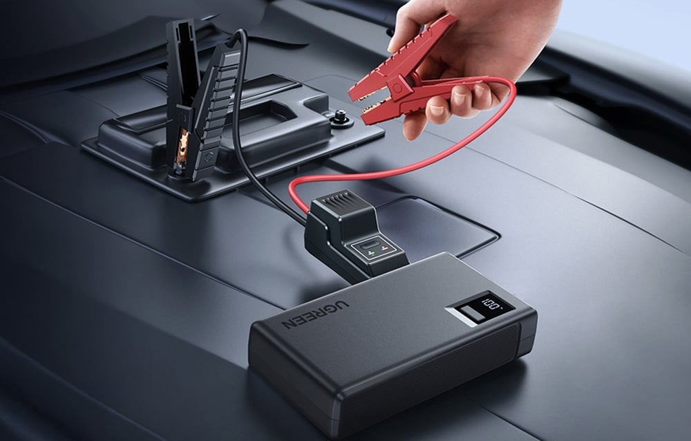 Egy kéz egy piros indítókábel-csipeszt tart egy autó akkumulátora felett. Egy fekete power bank 100%-ot mutat a kijelzőn.