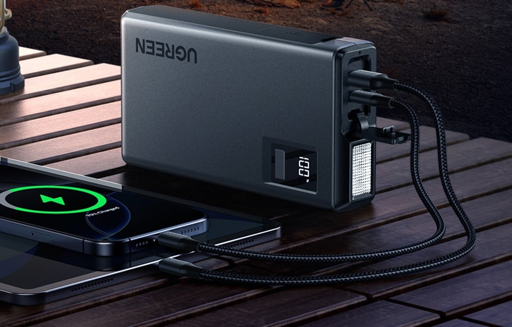 Egy fekete UGREEN power bank tölt egy telefont és egy tabletet. Kábelek kötik össze őket. Egy kijelző 100%-os töltést mutat.