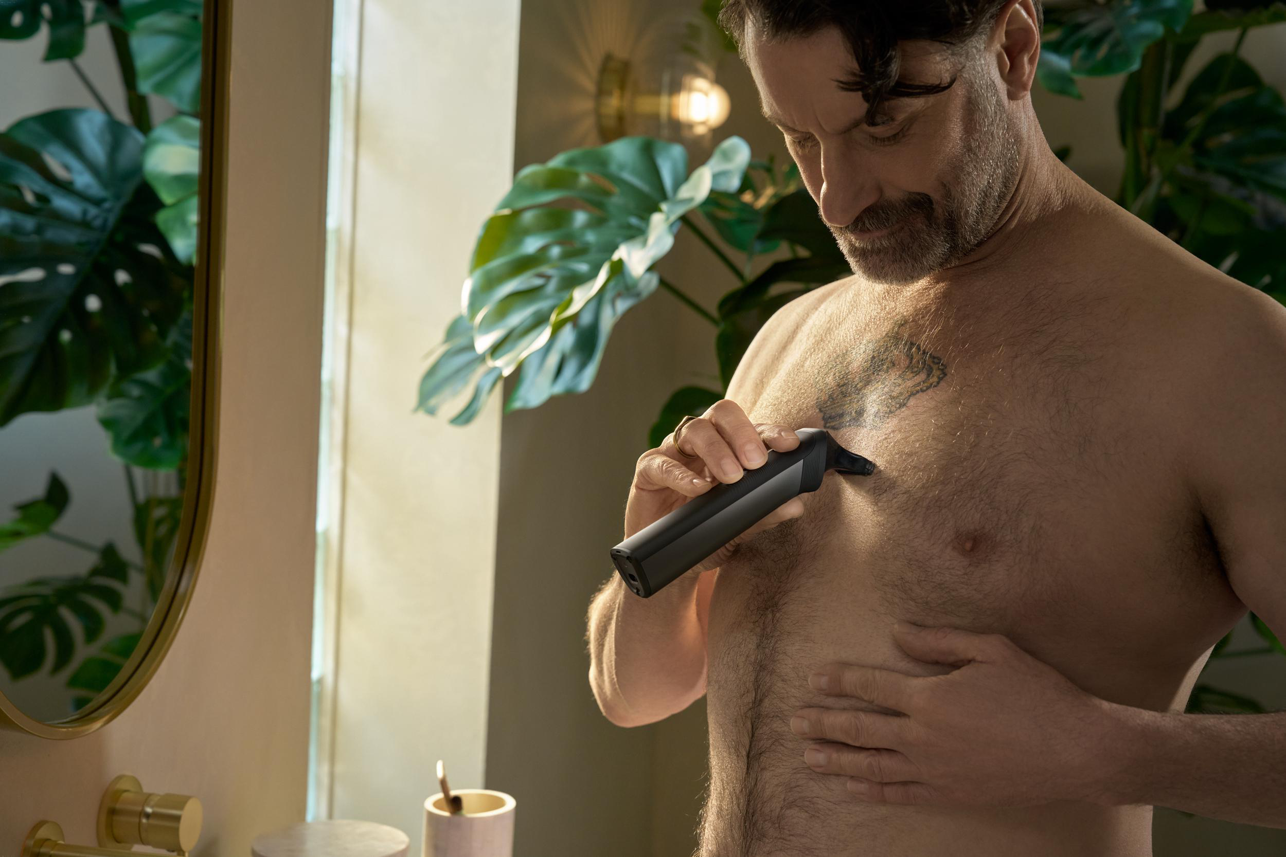 Ein Mann rasiert sich mit einem schwarzen Trimmer die Brusthaare. Grüne Blätter und ein Spiegel im Hintergrund.