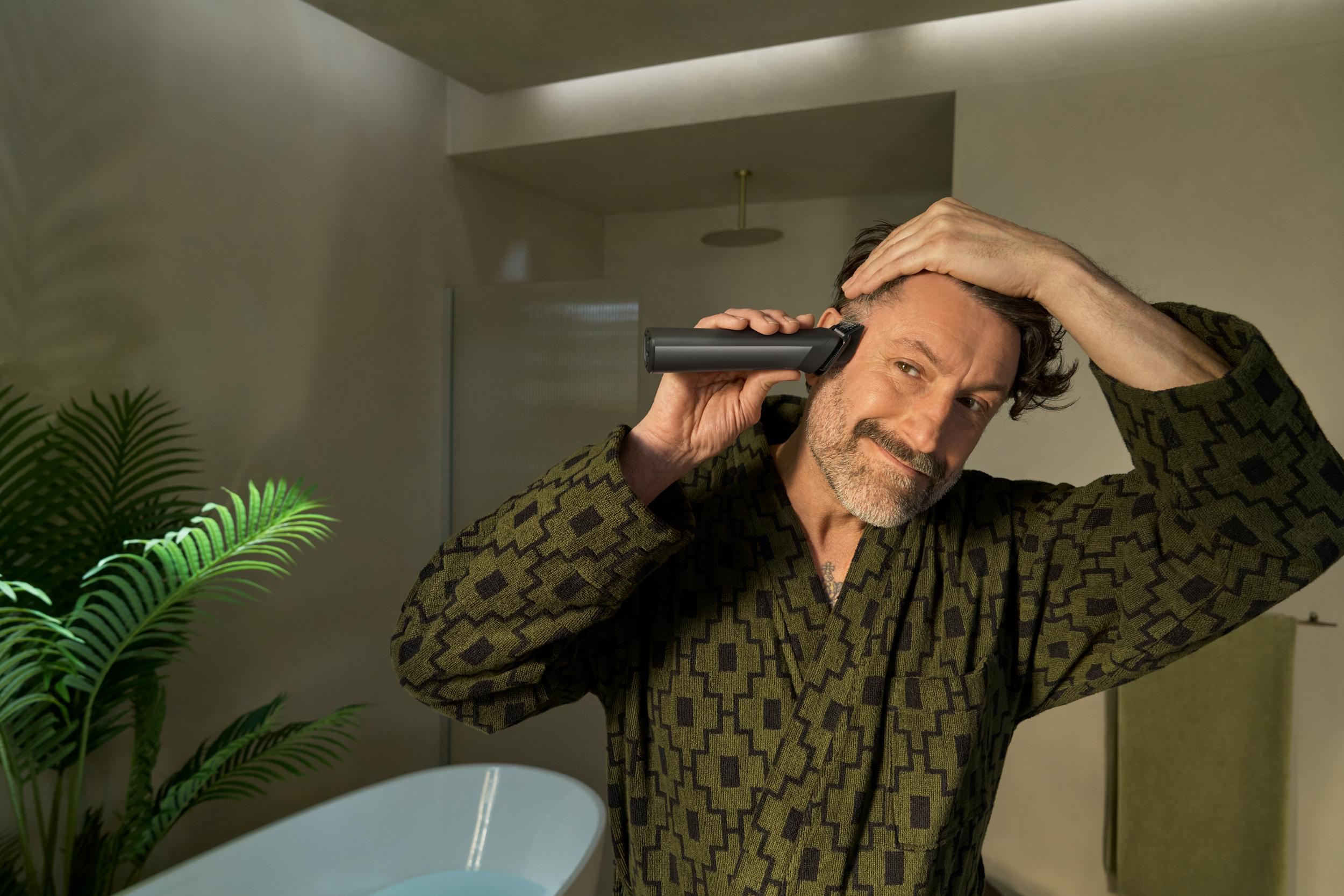 Ein Mann im Bademantel rasiert sich den Kopf. Er hält den Trimmer in der Nähe seines Kopfes. Im Hintergrund eine Badewanne und Pflanzen.