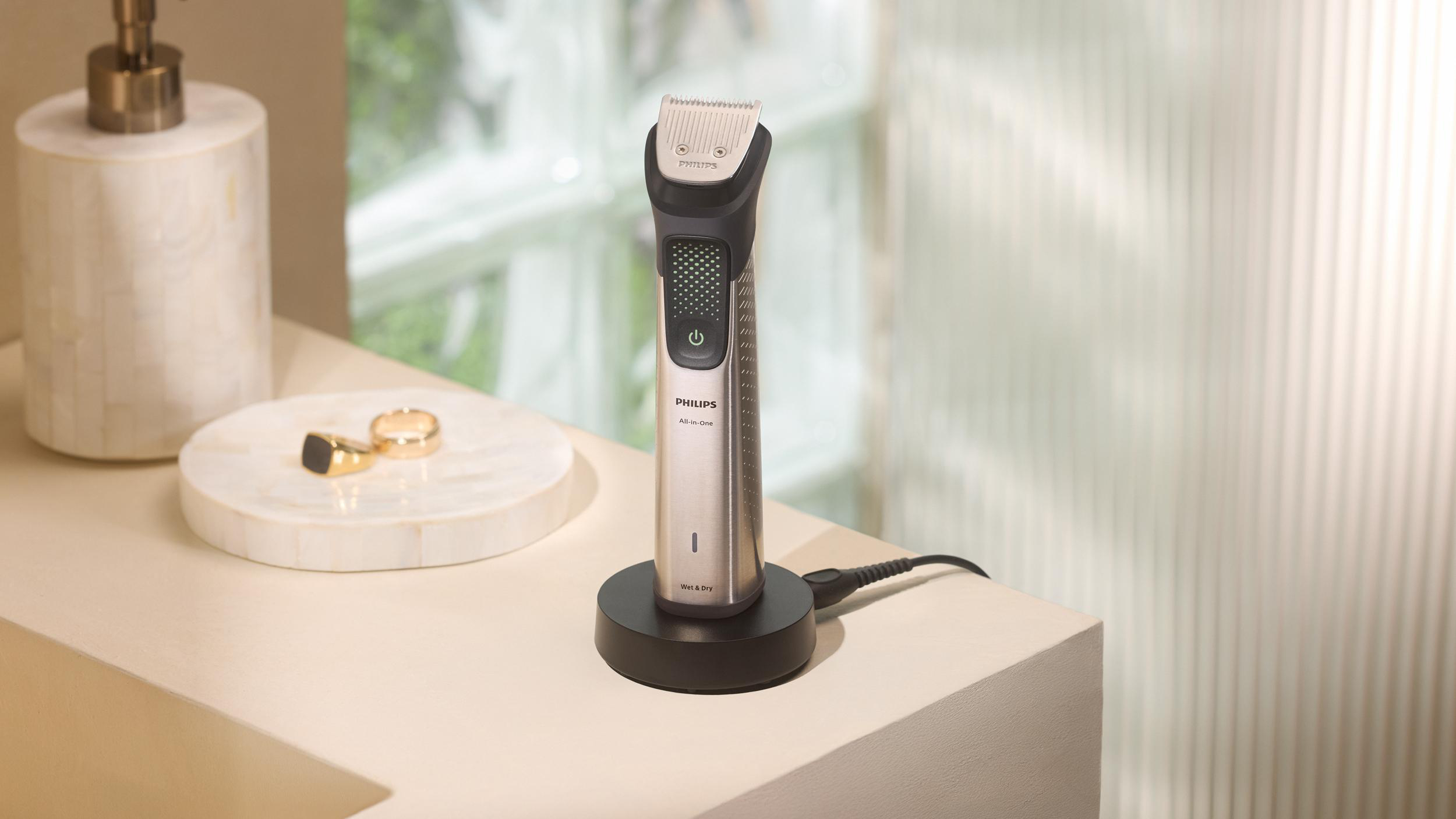 Ein Philips All-in-One Trimmer steht auf einer schwarzen Basis. Ein Seifenspender und Ringe sind in der Nähe. Der Hintergrund ist unscharf.