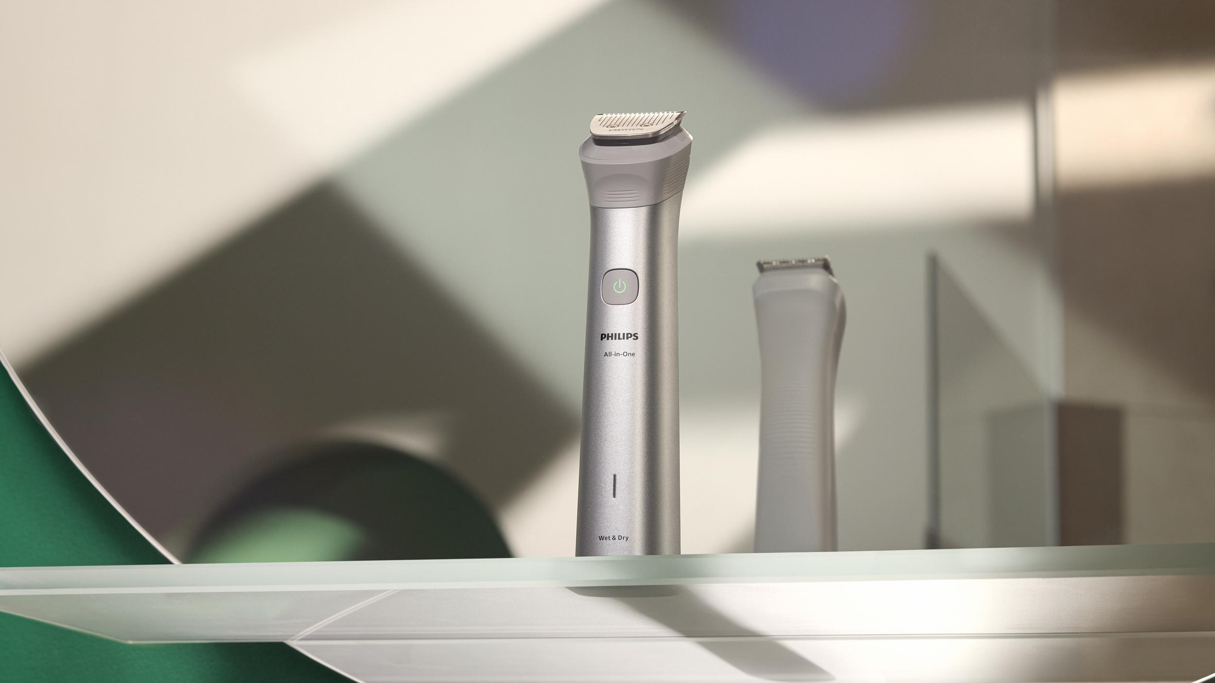 Ein Philips All-in-One Trimmer und ein Ersatzkopf liegen auf einem Regal. Der Trimmer ist silberfarben und hat einen grünen Knopf.