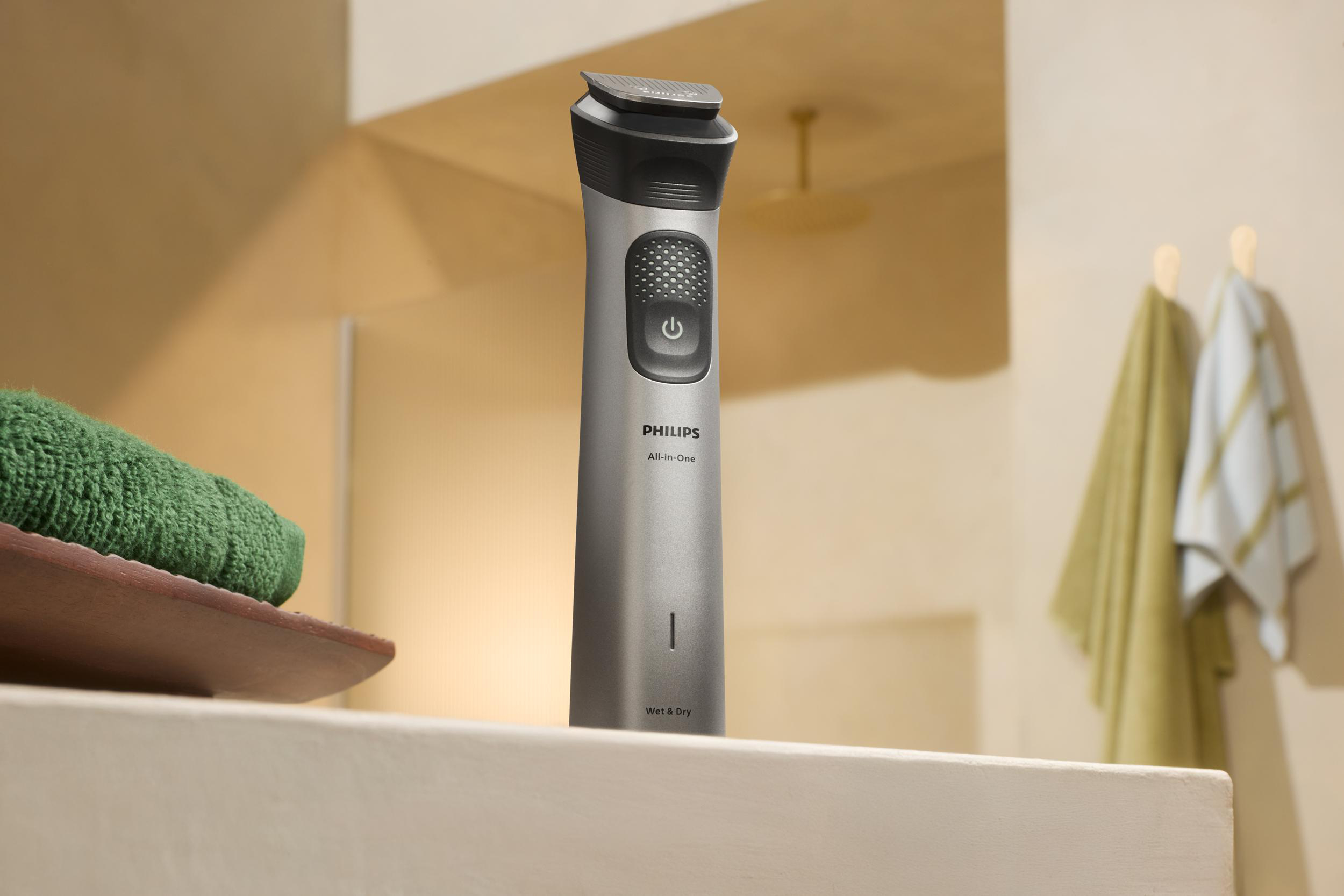 Ein Philips All-in-One-Trimmer steht aufrecht. Ein grünes Handtuch liegt auf einer Holzablage. Zwei Handtücher hängen im Hintergrund.