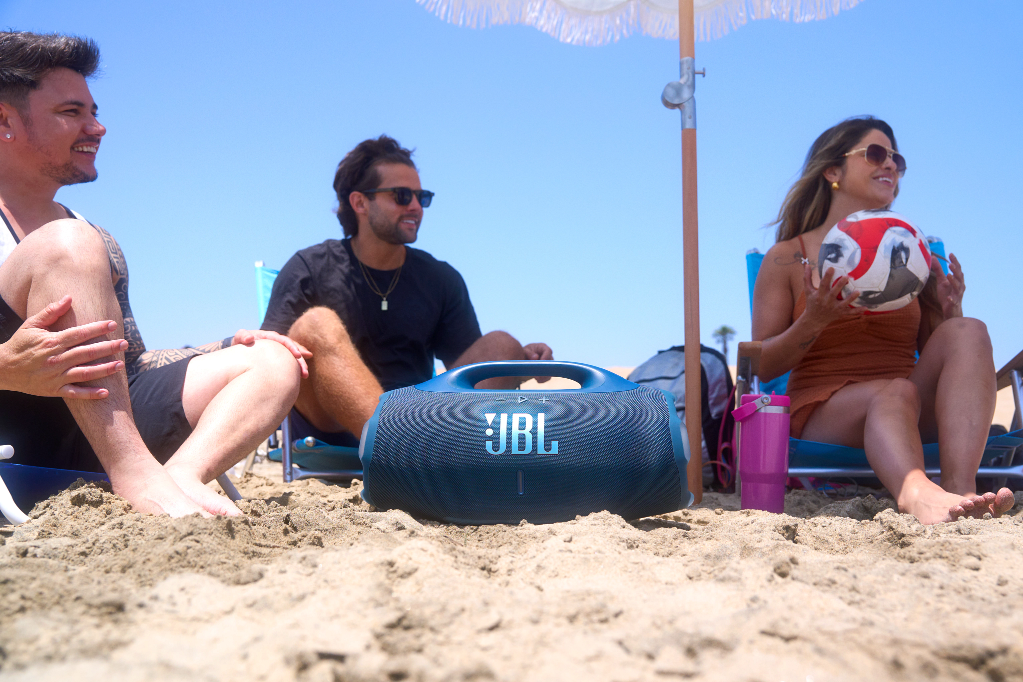Drei Personen am Strand, ein JBL-Lautsprecher davor. Eine Frau hält einen Ball. Sonnenschirm spendet Schatten. Blauer Himmel im Hintergrund.