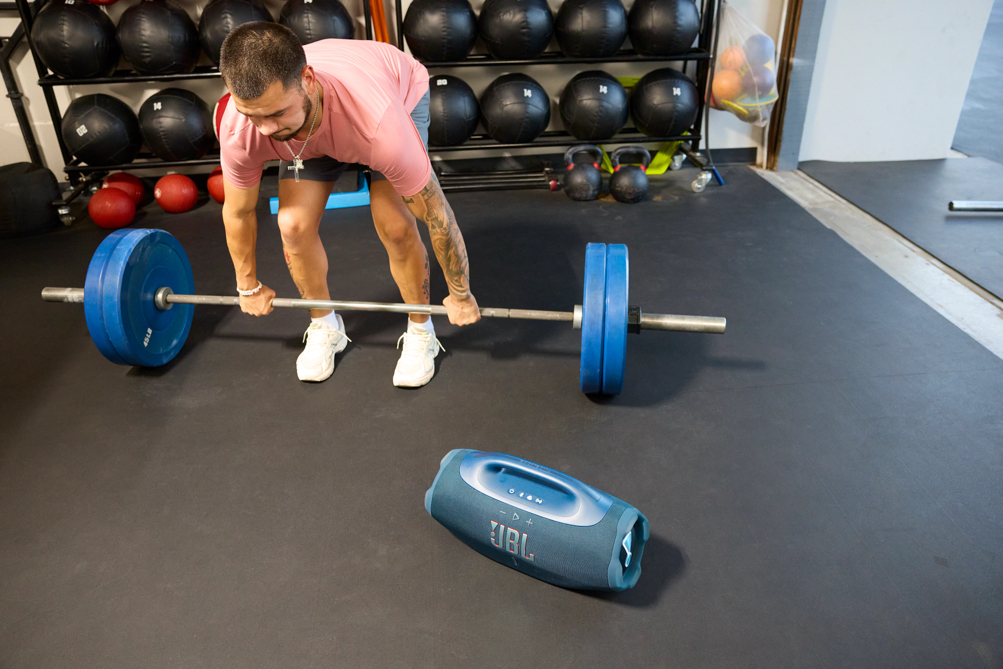 Mann in rosa Hemd hebt Langhantel mit blauen Gewichten. Ein JBL-Lautsprecher steht daneben auf dem Boden des Fitnessstudios. Fitnessgeräte im Hintergrund.