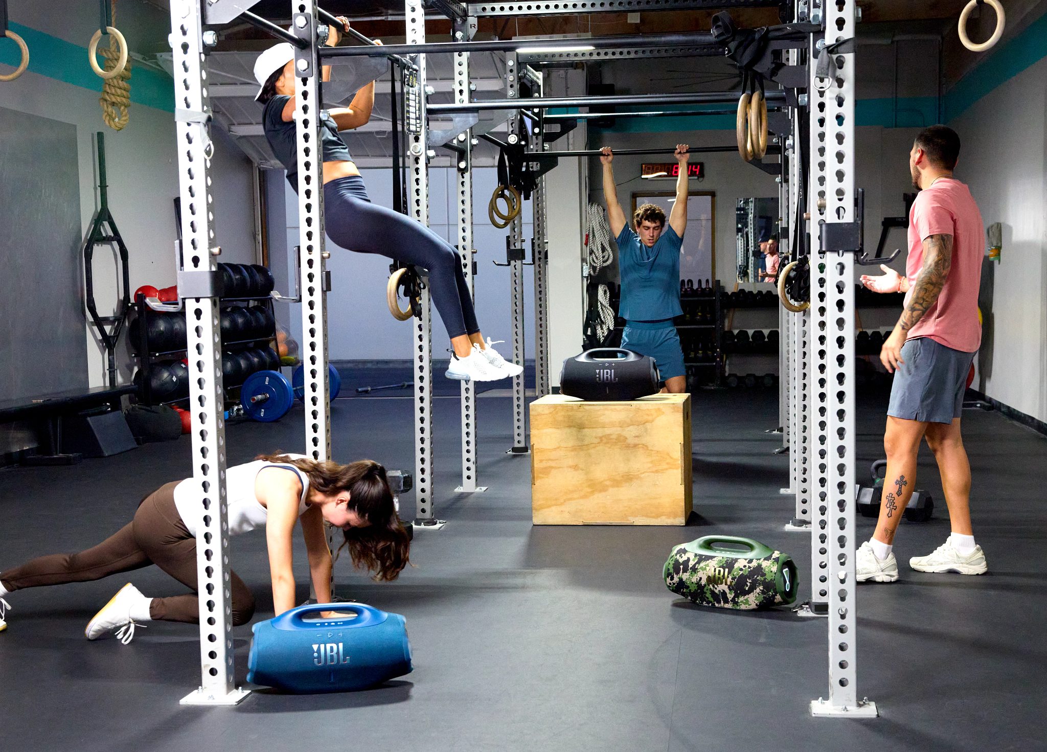 Leute trainieren in einem Fitnessstudio. Eine Person macht Klimmzüge. Andere trainieren. JBL-Lautsprecher und Holzkiste vorhanden.