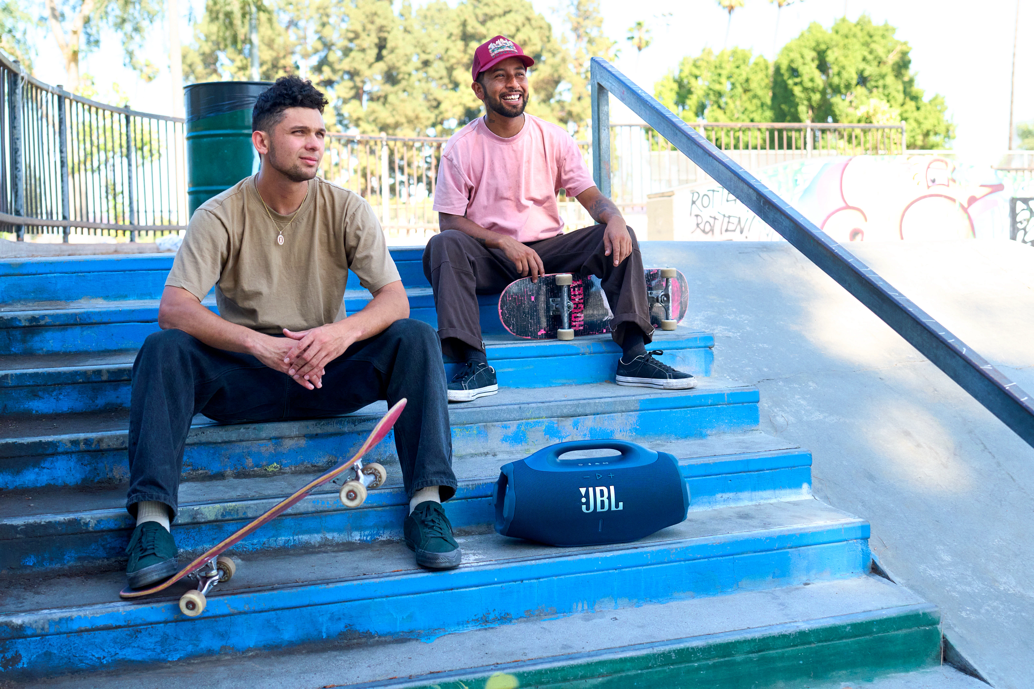Zwei Männer sitzen auf blauen Treppen mit Skateboards und einem JBL-Lautsprecher. Einer trägt ein pinkes Hemd und eine Kappe, der andere ein beiges Hemd.
