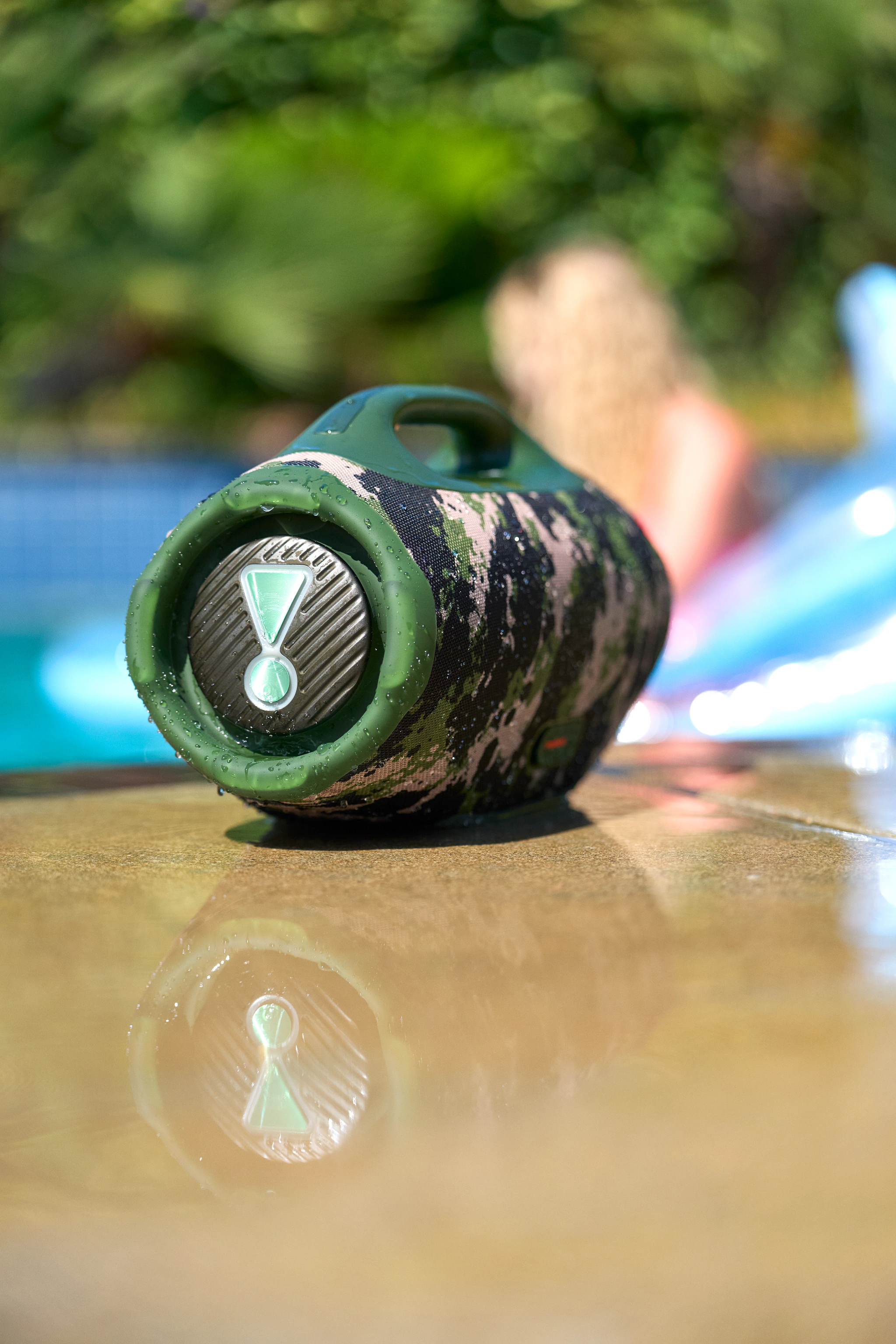 Ein Camouflage-Lautsprecher steht auf einer Oberfläche. Der Hintergrund hat einen verschwommenen Pool und eine Person.