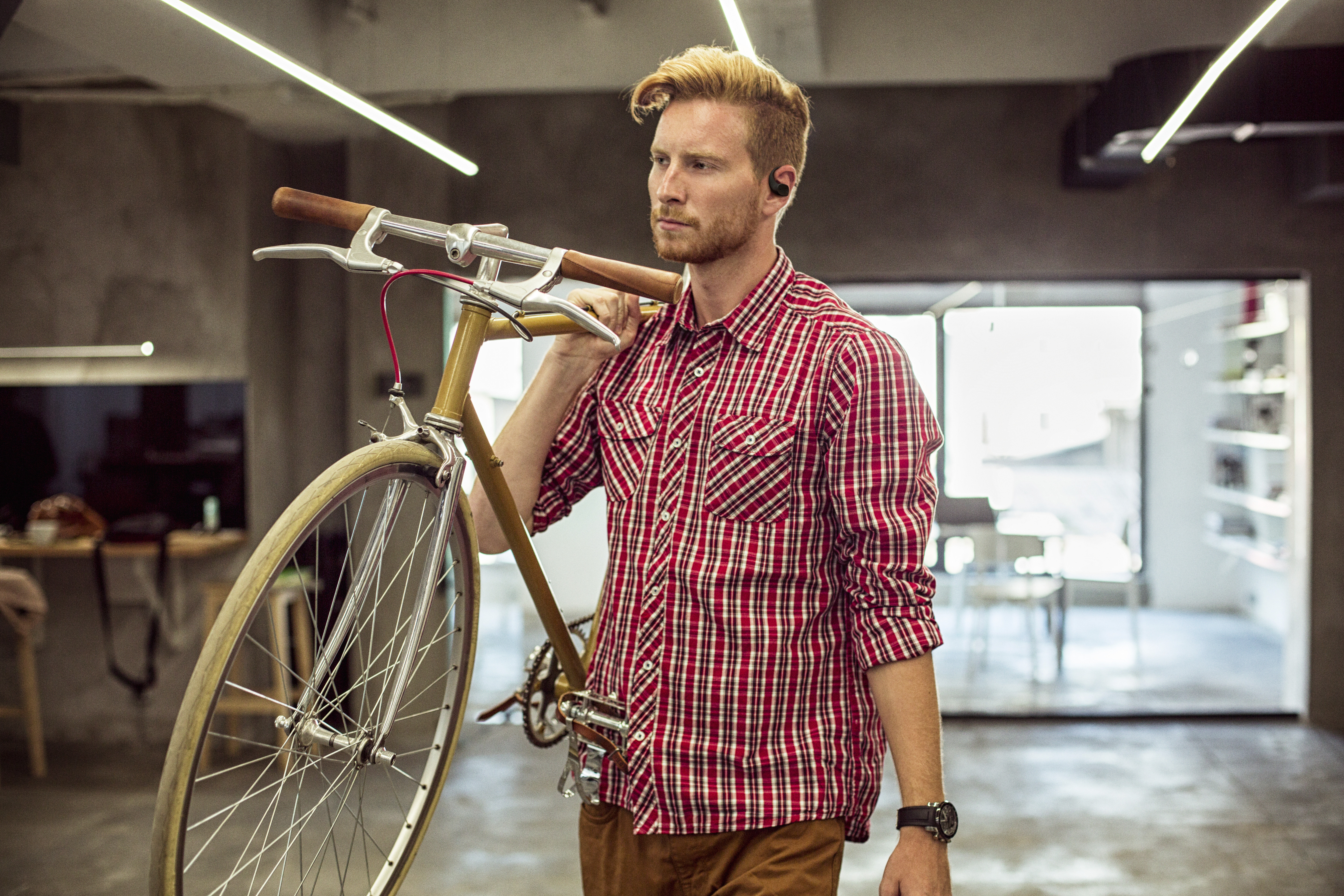 Un homme en chemise à carreaux rouge porte un vélo. Il a des écouteurs et une montre. Le cadre semble être un bureau ou un magasin.