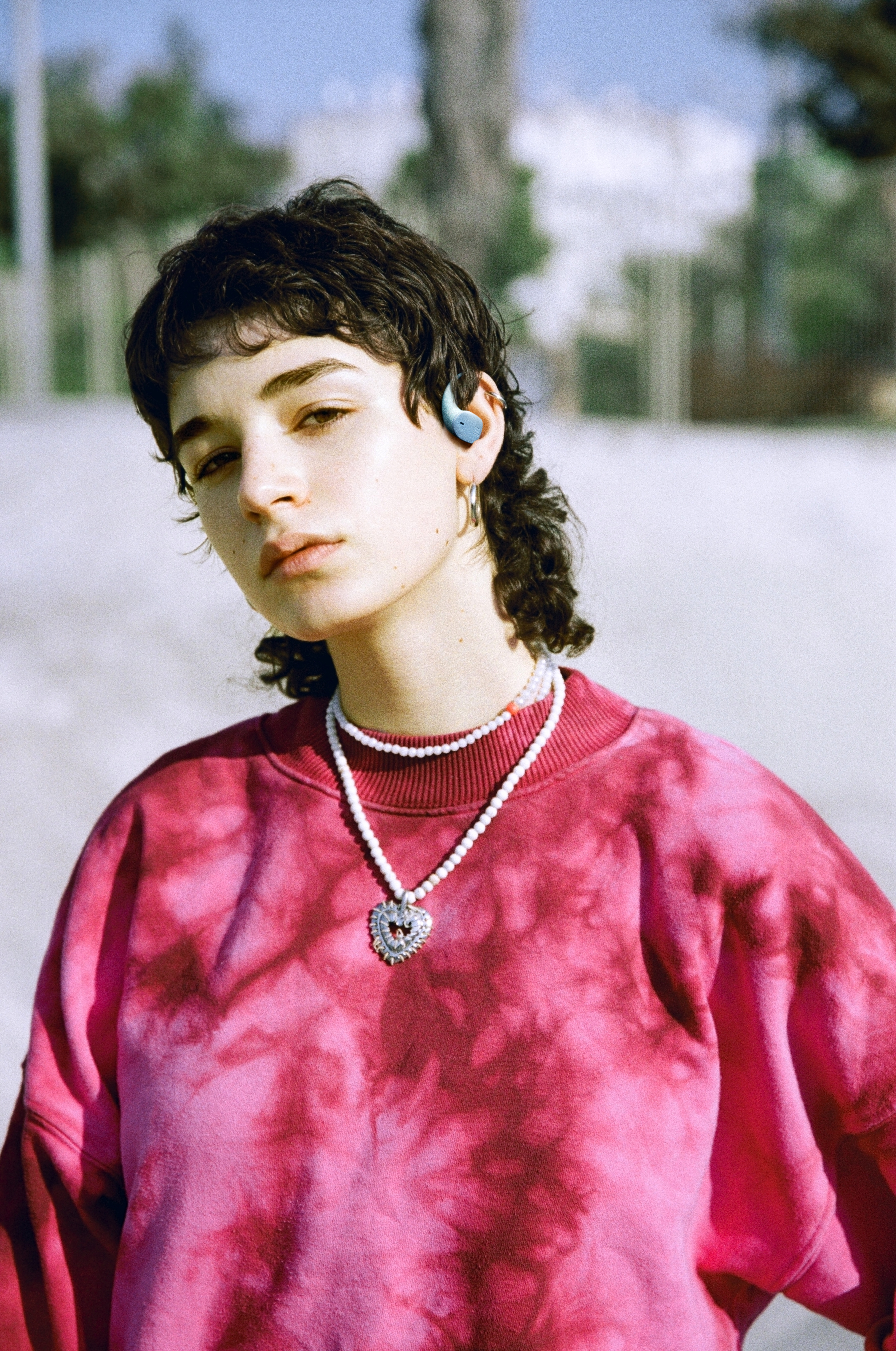 Eine Person mit dunklen Haaren, die ein rosa Batik-Shirt, eine Perlenkette mit Herzanhänger und einen blauen Ohrhörer trägt.