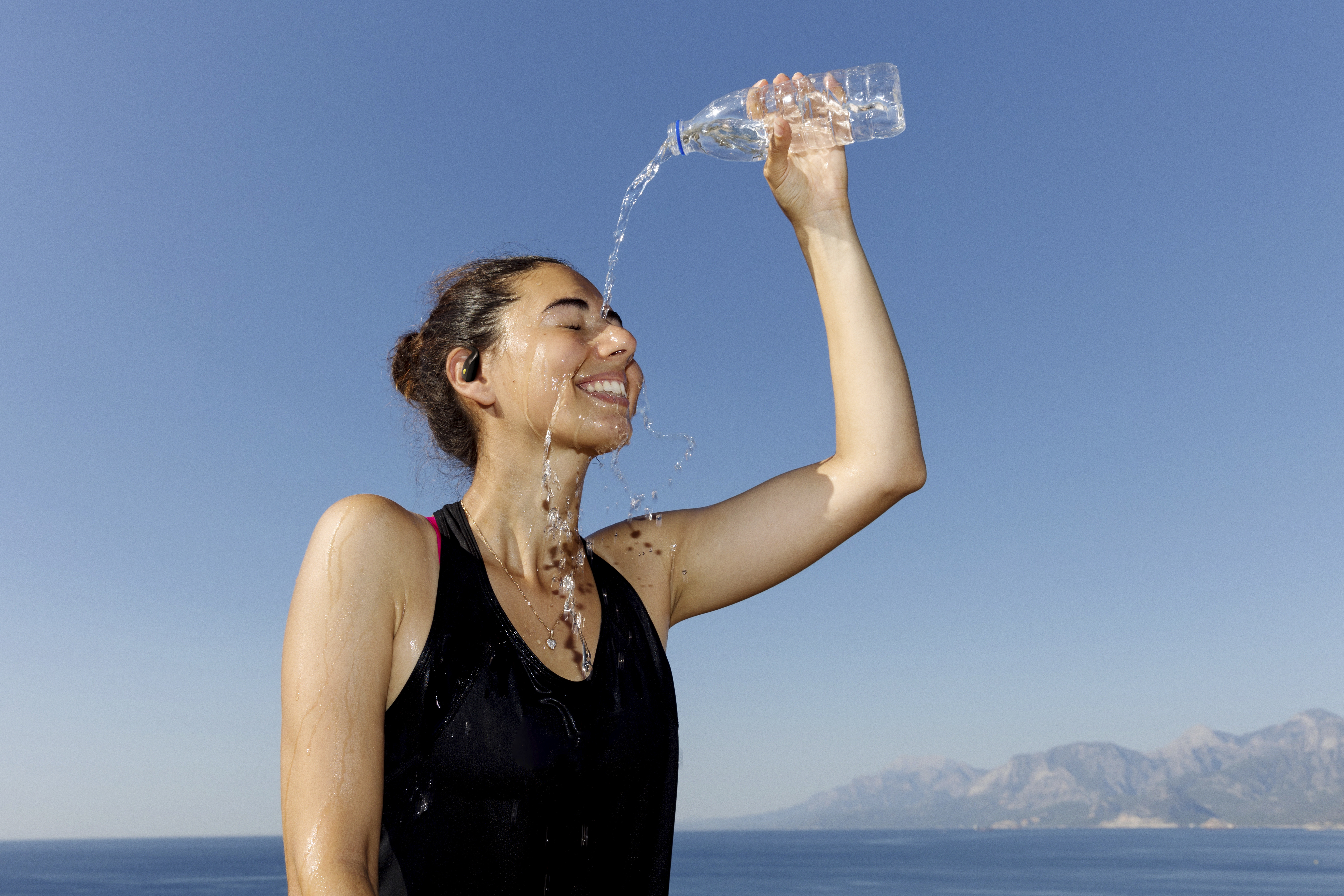 Eine Frau gießt sich Wasser aus einer Flasche ins Gesicht. Sie lächelt, trägt ein schwarzes Top, im Hintergrund Meer und Berge.