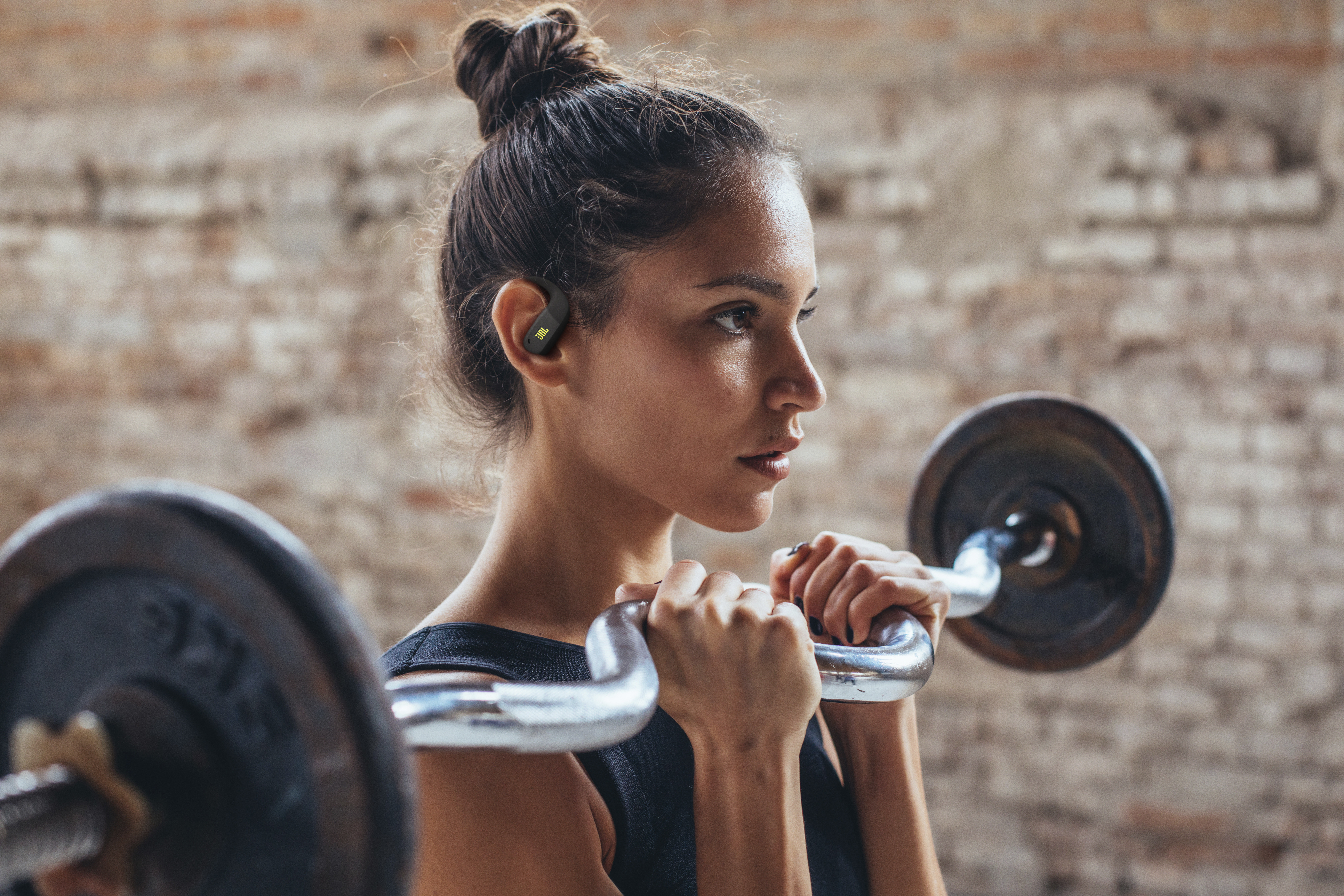 Frau im Fitnessstudio mit Langhantel, trägt Ohrhörer. Braune Haare hochgesteckt, schwarzes Oberteil, seitlich blickend. Ziegelwand im Hintergrund.