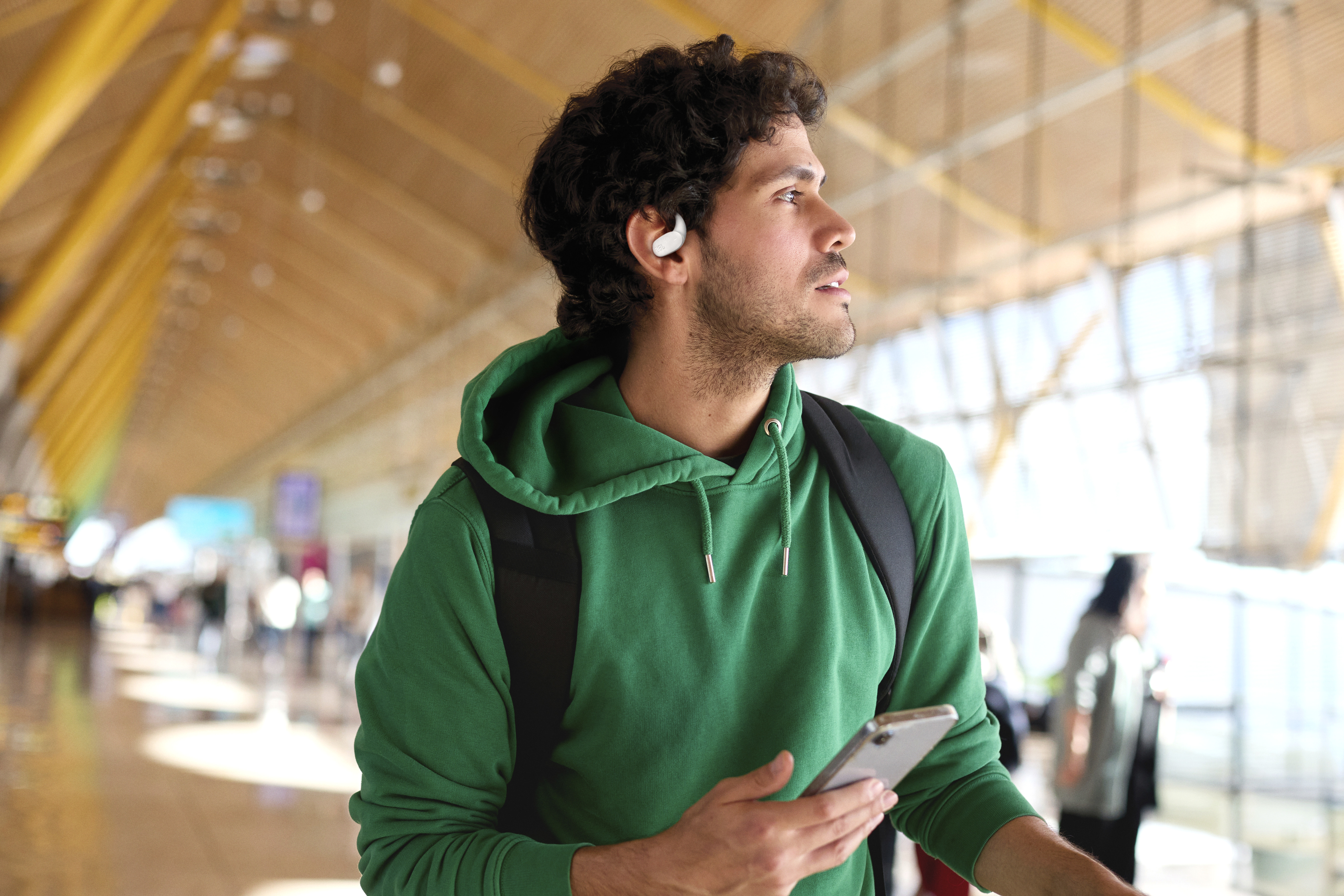 Mann im grünen Kapuzenpullover am Flughafen, Handy haltend, Ohrhörer tragend. Rucksack auf. Blick nach rechts.
