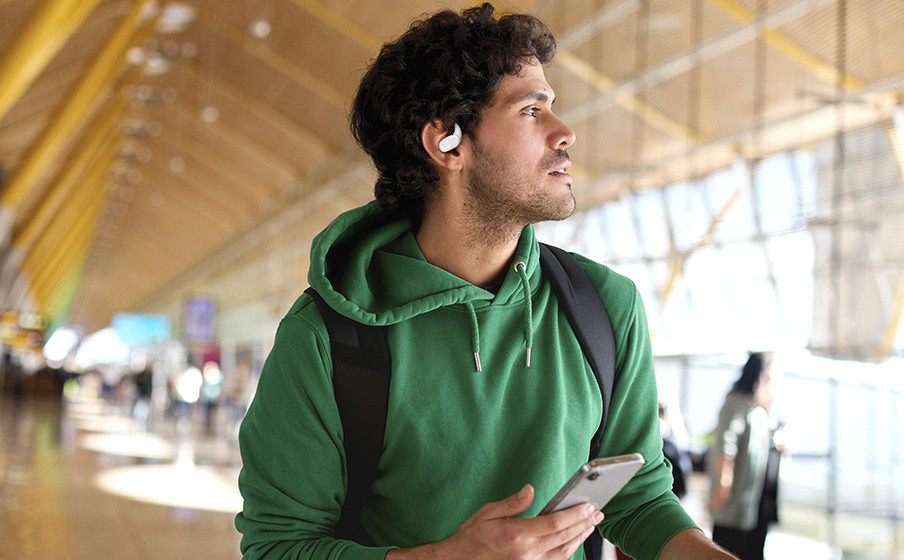 Mann mit grünem Kapuzenpullover und Rucksack mit Ohrhörer in einem Flughafen, der ein Telefon hält. Verschwommener Hintergrund mit Menschen und Architektur.