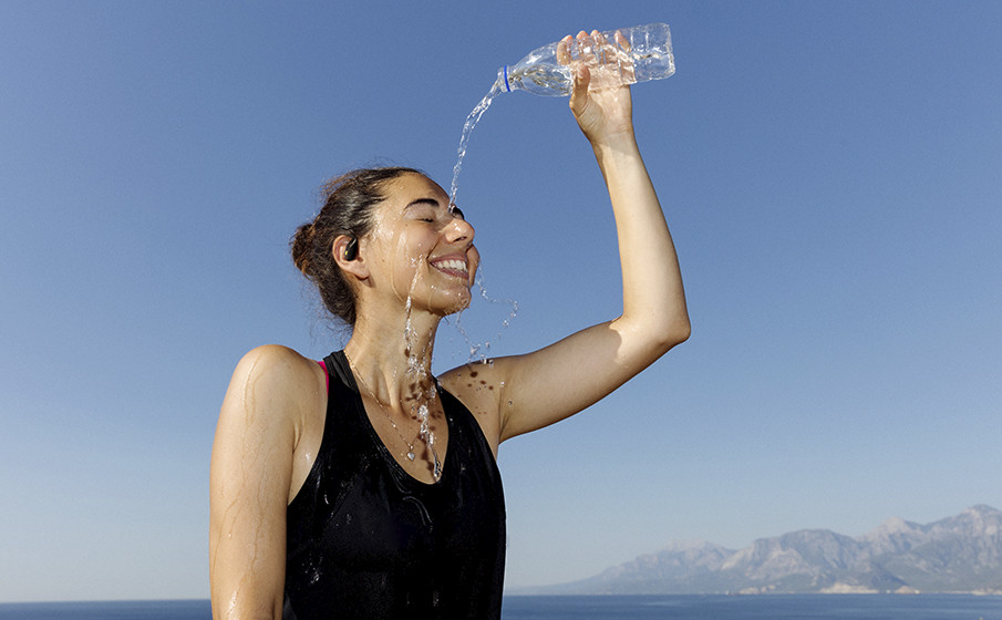 Frau gießt sich Wasser aus einer Flasche über den Kopf. Sie trägt ein schwarzes Oberteil und lächelt. Klarer Himmel und Berge im Hintergrund.