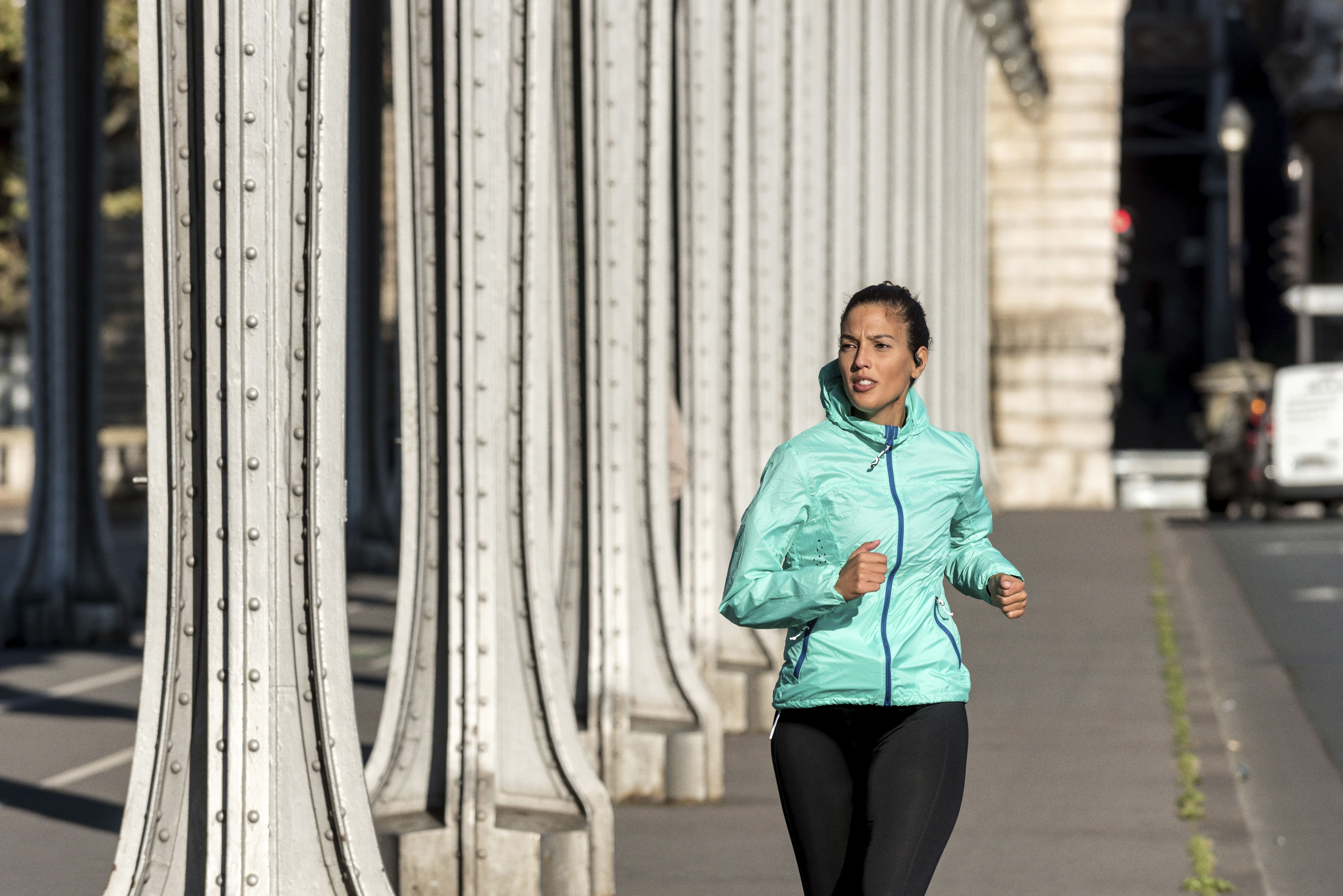 Eine Frau läuft auf einer Brücke, trägt eine türkisgrüne Jacke und schwarze Leggings. Die Brücke hat Metallsäulen. Im Hintergrund ein Fahrzeug.