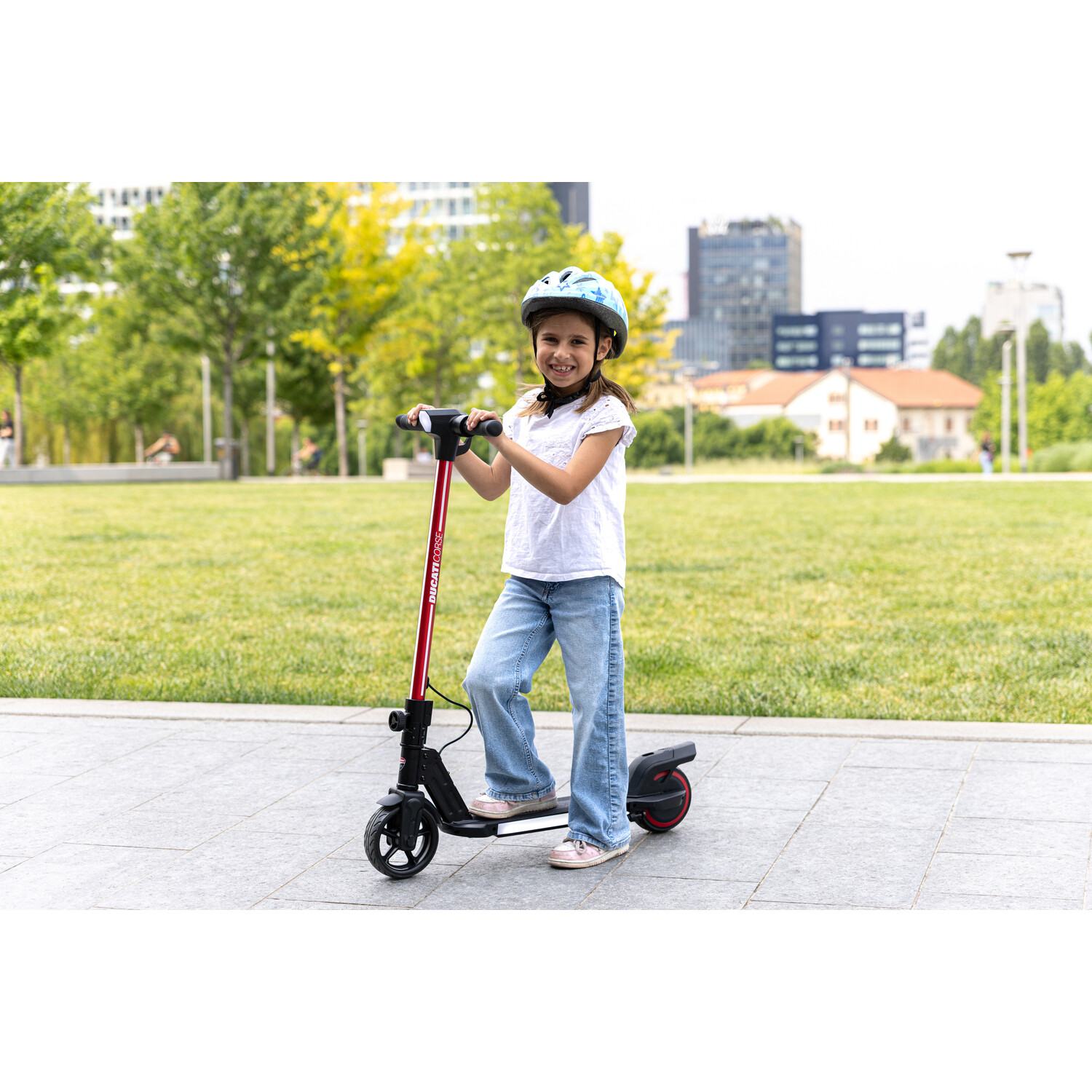 Een meisje met een helm staat op een elektrische scooter. Ze glimlacht in een park. Gebouwen en bomen op de achtergrond.
