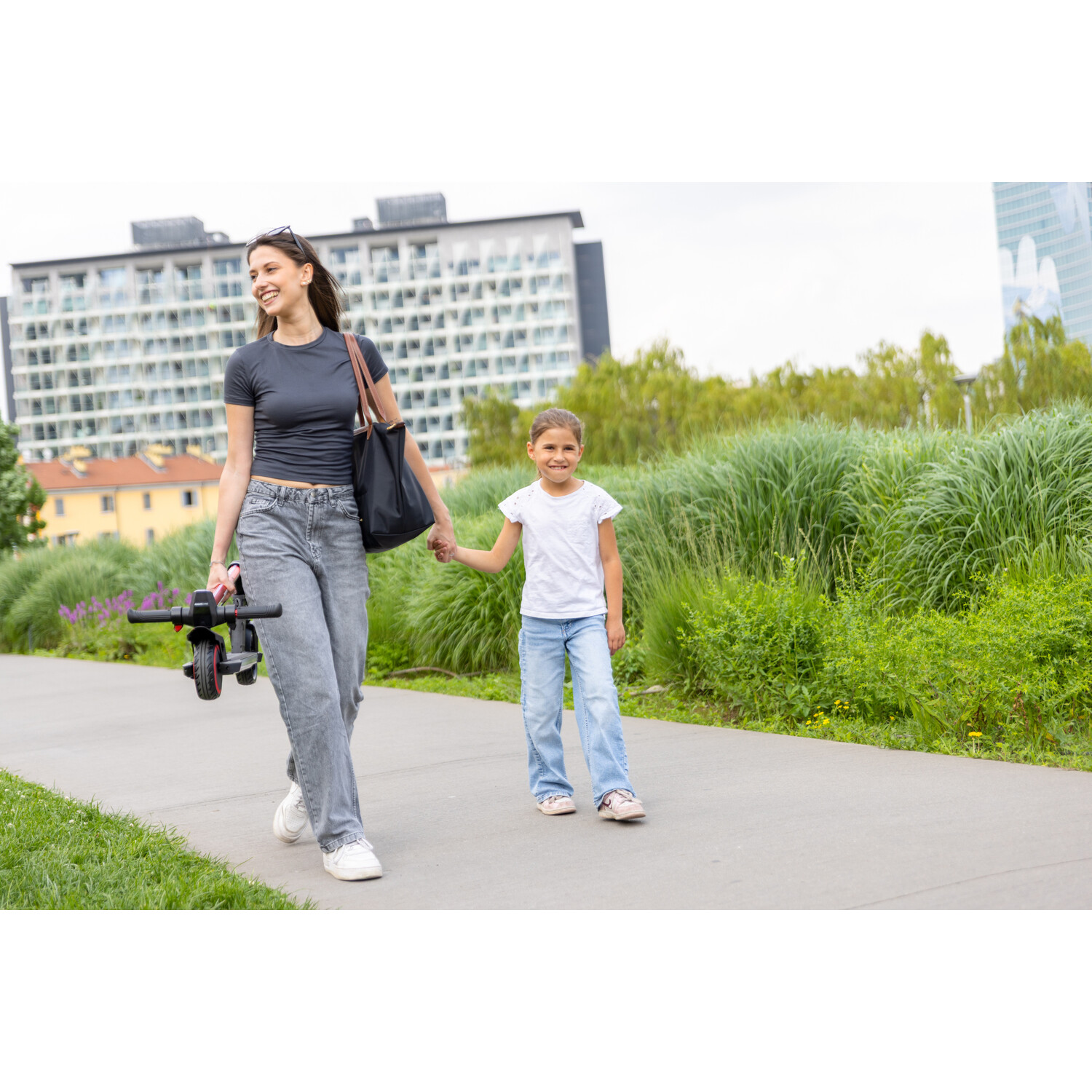 Een vrouw en een meisje lopen hand in hand op een pad. De vrouw draagt een scooter. Groen gras en gebouwen op de achtergrond.