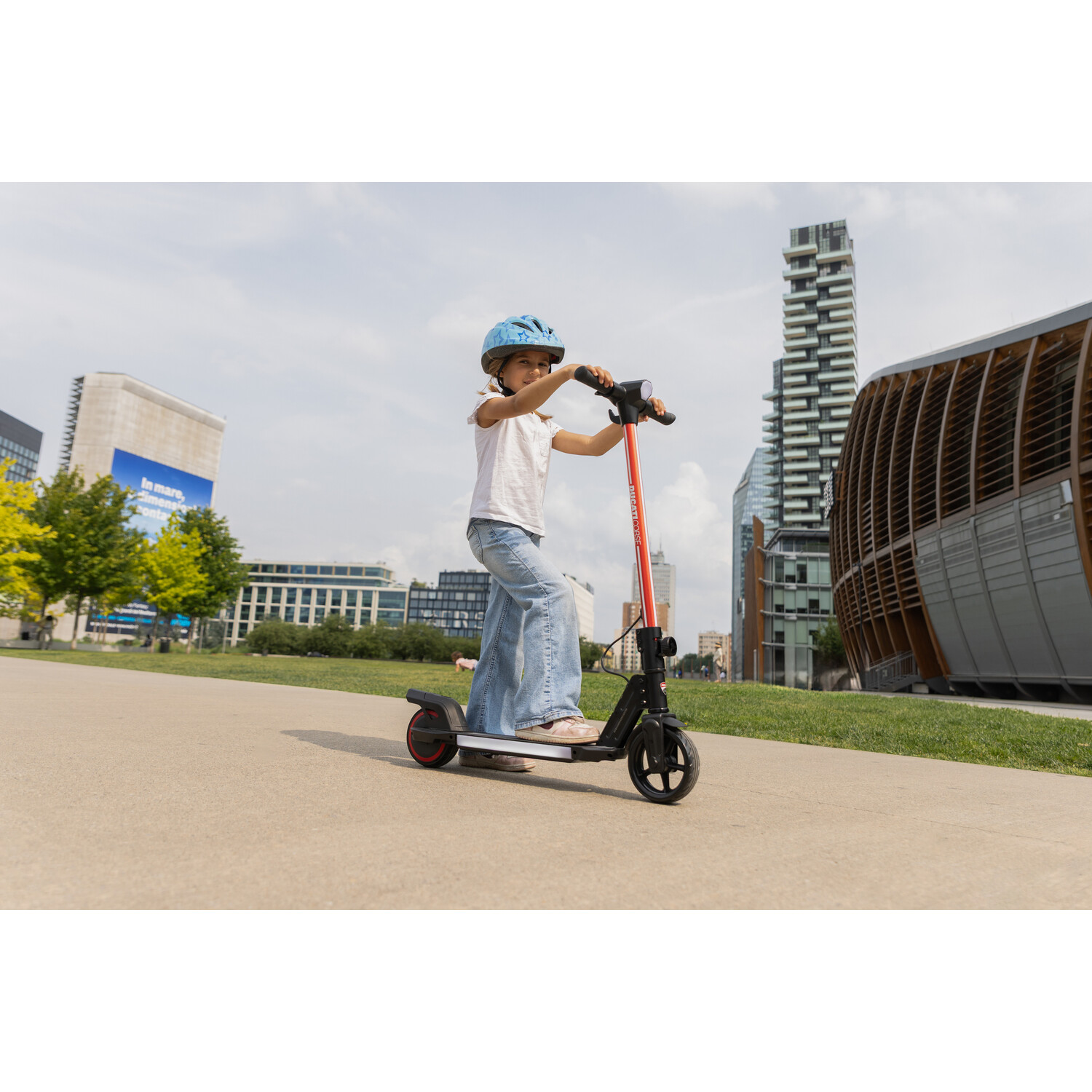 Een jong meisje rijdt op een rood-zwarte scooter op een pad. Ze draagt een helm en een spijkerbroek. Gebouwen en bomen op de achtergrond.