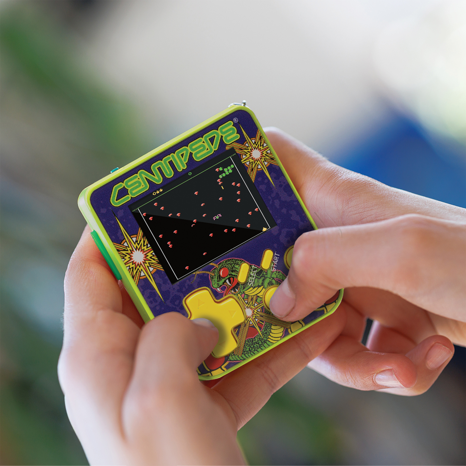 Mains tenant un mini jeu d'arcade. L'écran affiche un jeu Centipede avec des éléments rouges et verts. Le jeu est violet et vert.