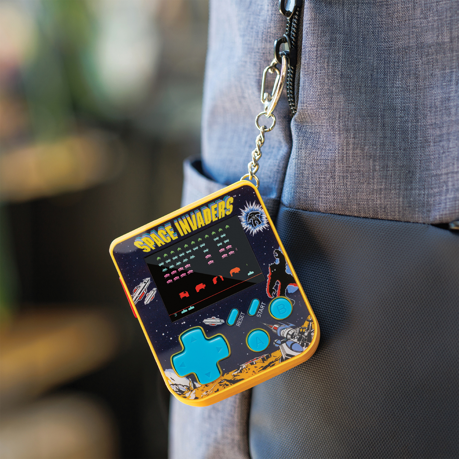 Un jeu portable Space Invaders, cadre jaune, boutons bleus, suspendu à une chaîne sur un sac à dos gris. L'écran montre des extraterrestres et des vaisseaux.