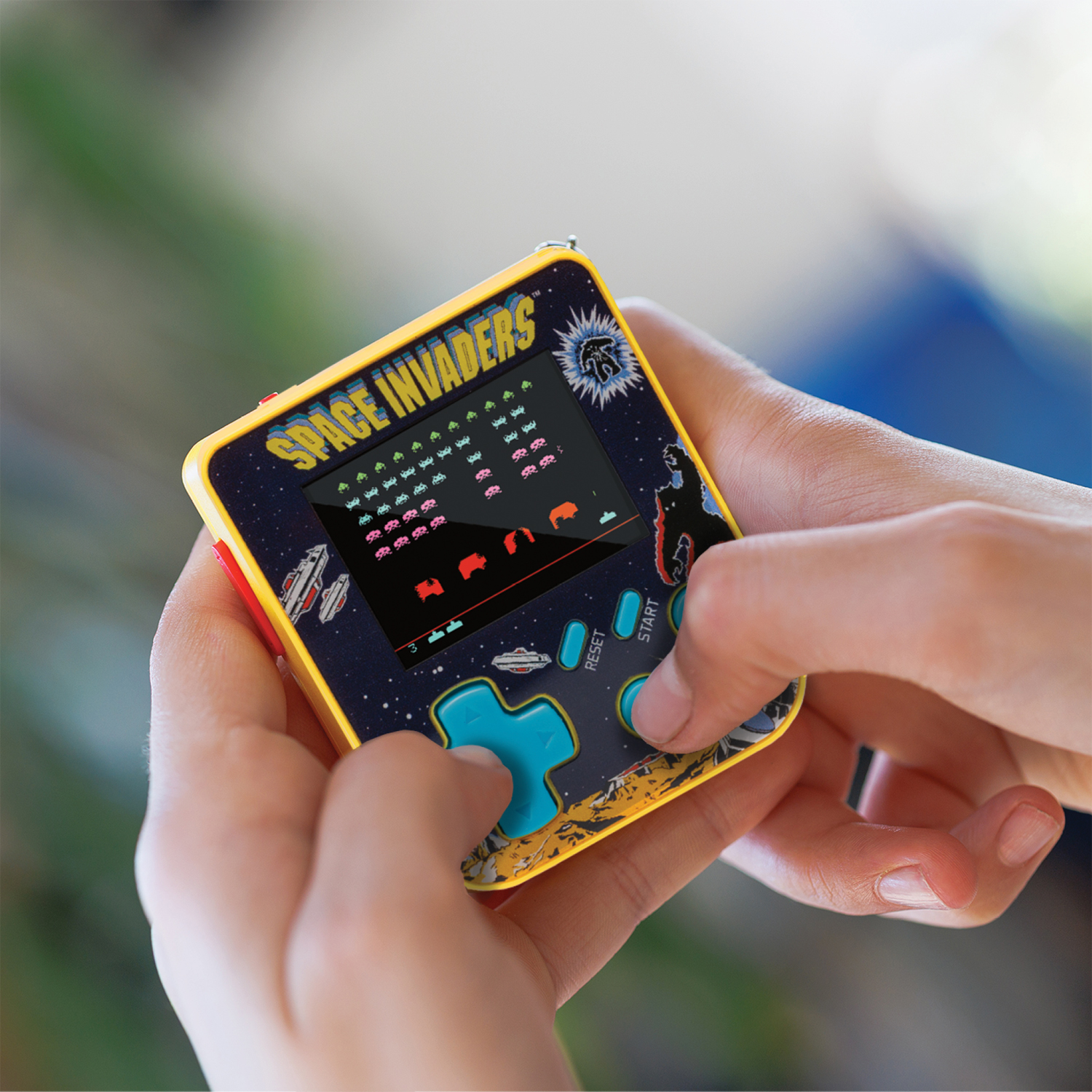 Mains tenant un jeu portable Space Invaders. L'écran affiche le jeu en cours. L'appareil a des boutons bleus et un bord jaune.