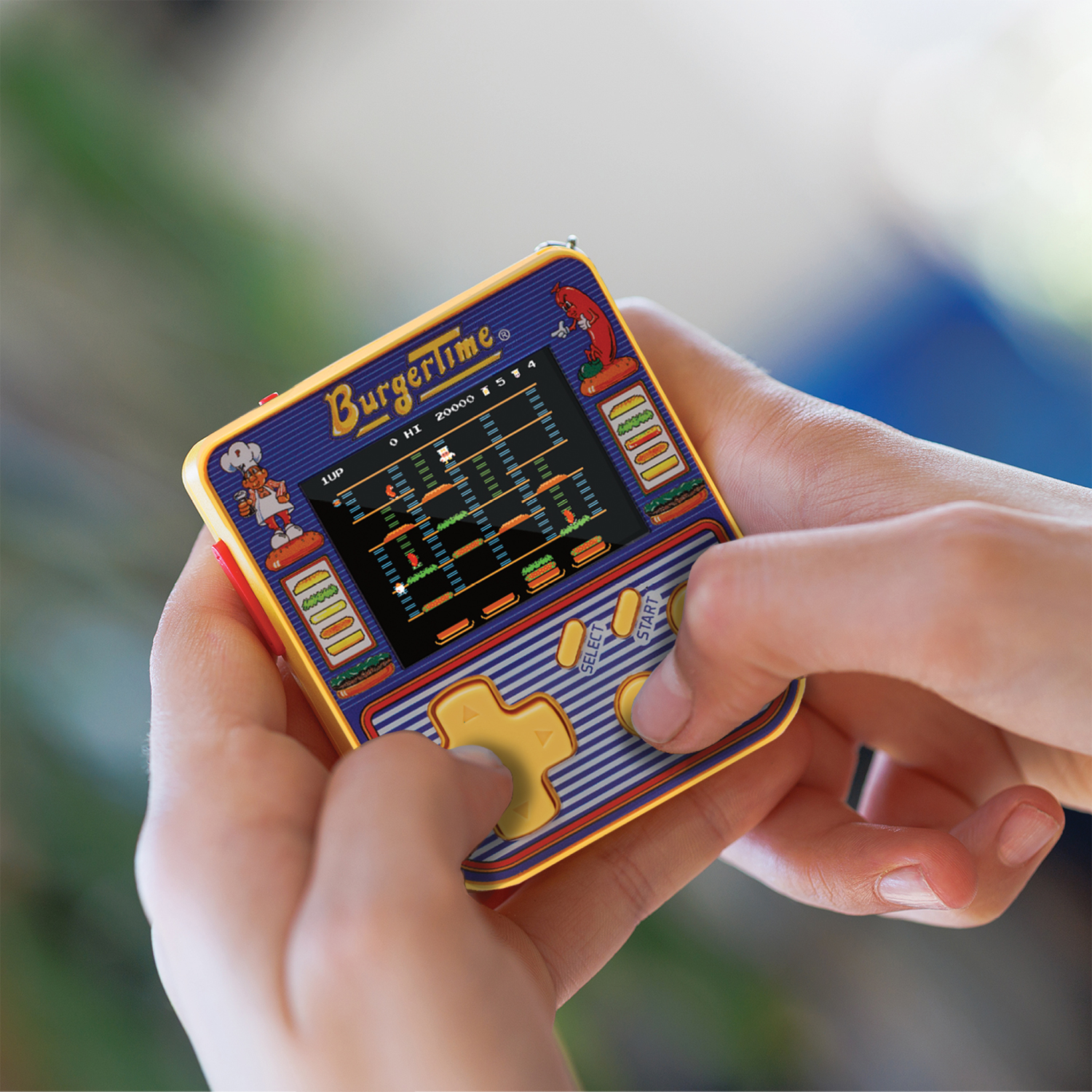 Mains tenant une console portable jaune BurgerTime. L'écran montre le jeu. Le fond est flou.