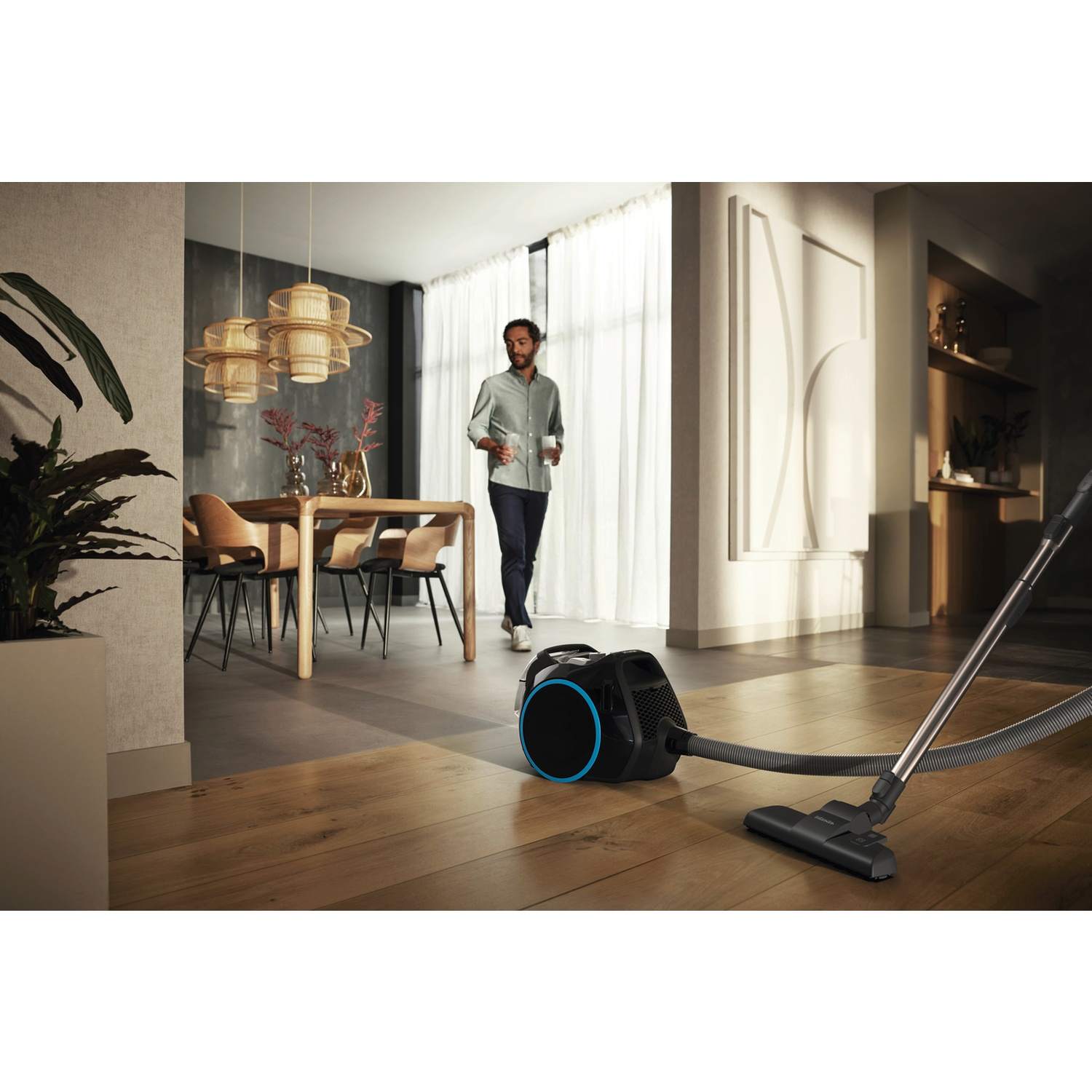A man walks past a vacuum cleaner on wooden floor. A table and chairs are in the background. The room is well lit.