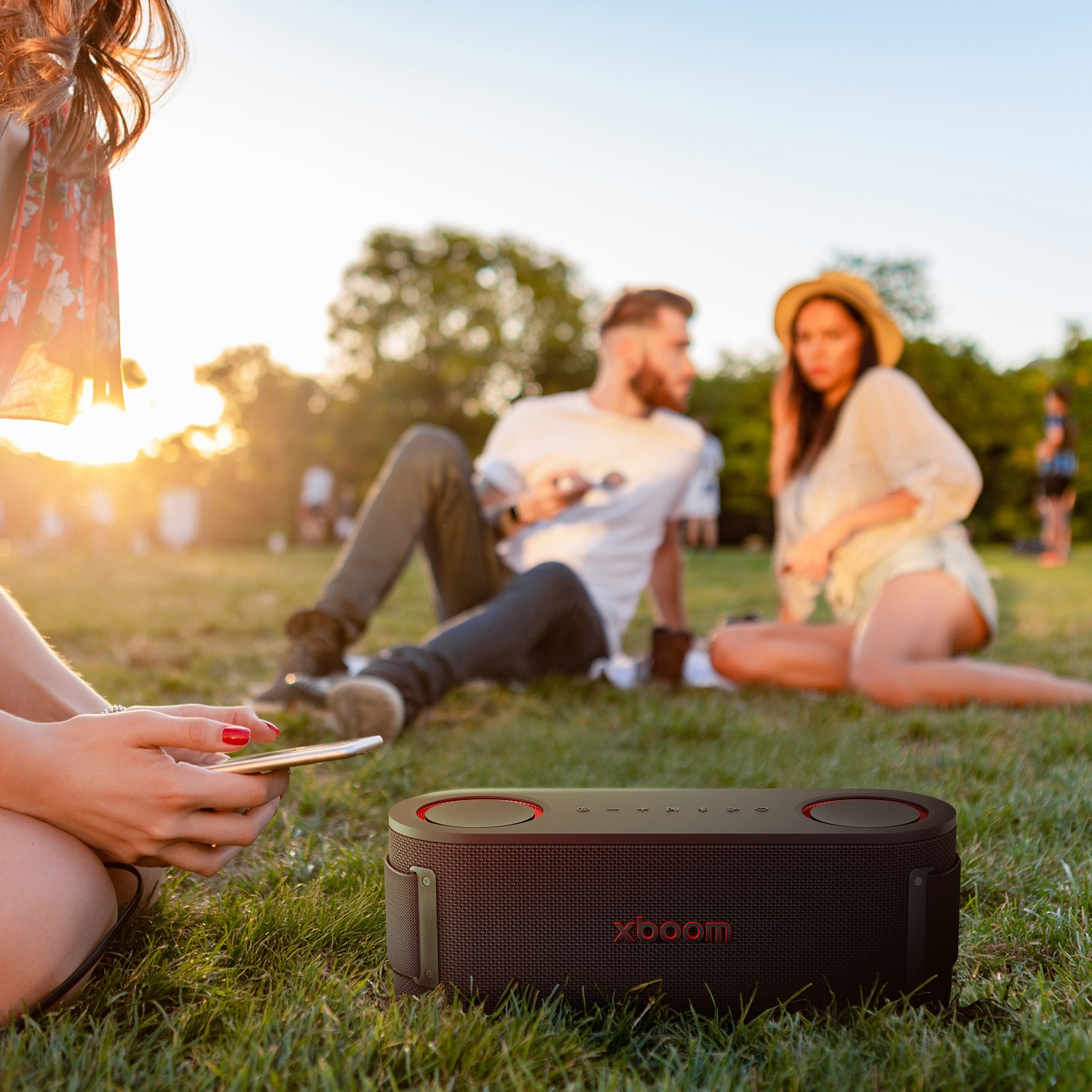 Persone in un parco che ascoltano musica. Un altoparlante nero è in primo piano. Due persone sono sedute sull'erba. Una persona tiene un telefono.