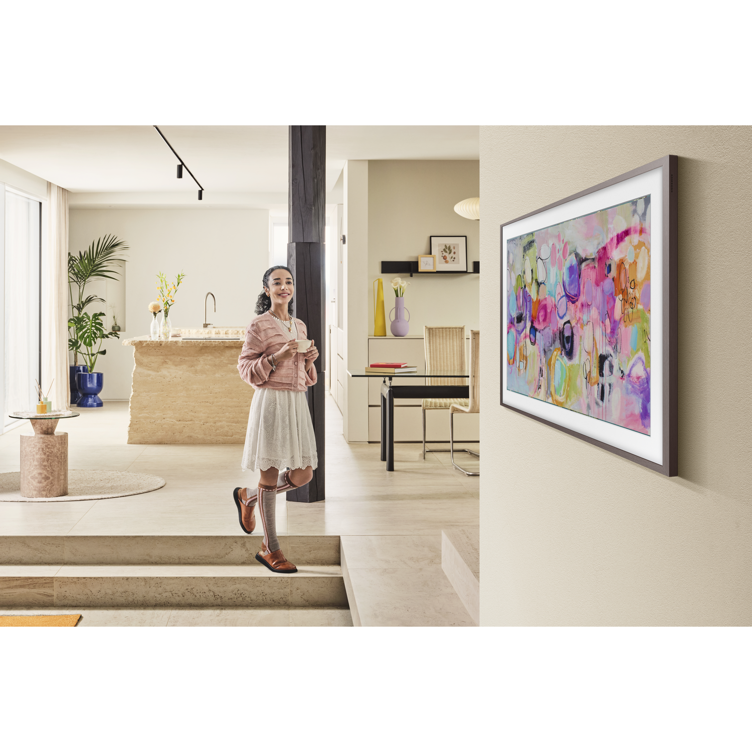 A woman in a pink sweater and white skirt stands near a painting. The room has modern decor, with a kitchen and a dining area.