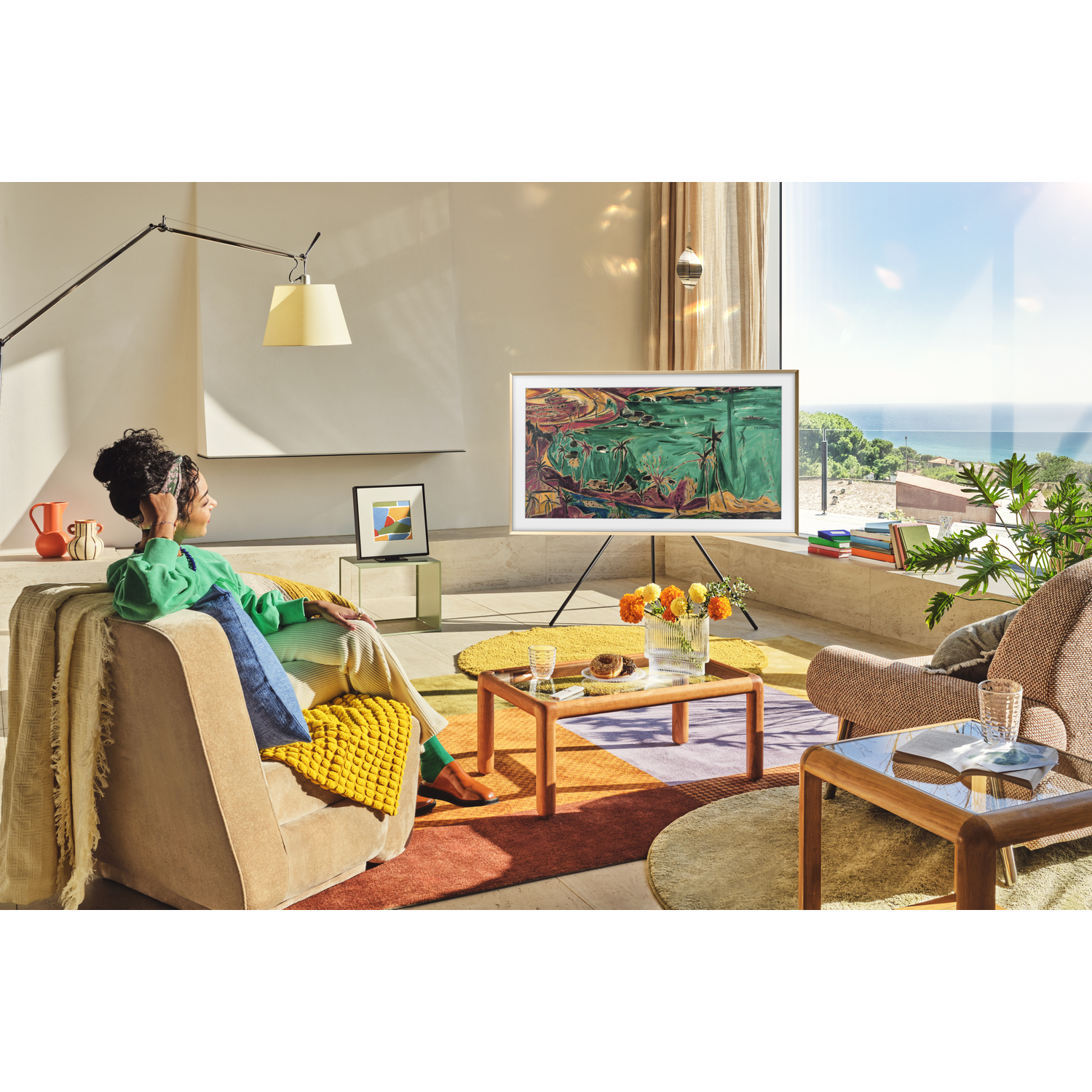 Woman sits on a couch, looking at TV. A colorful rug, coffee tables, and a view of the ocean are in the background.