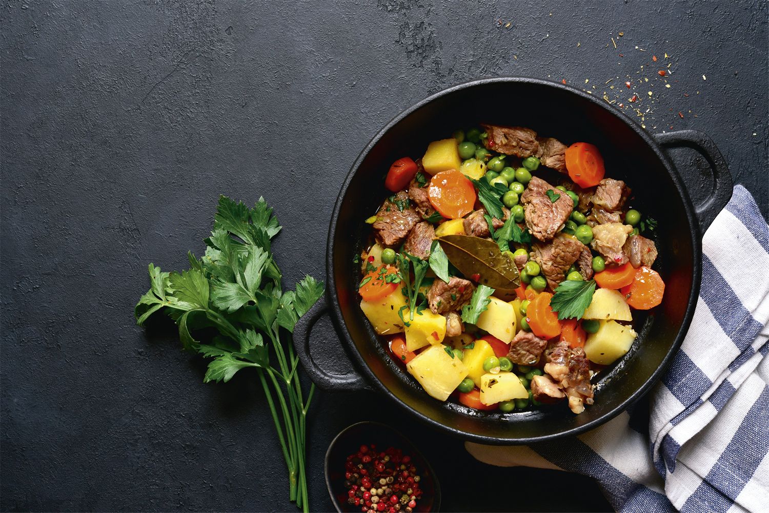 Estofado en una olla negra con carne, zanahorias, guisantes y patatas. Perejil y un cuenco pequeño de granos de pimienta.