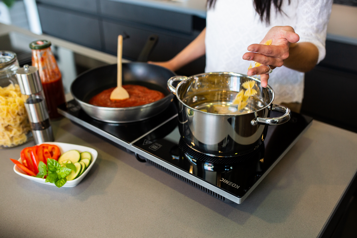 Una mujer cocina pasta. Se dejan caer fideos farfalle en una olla. La salsa hierve a fuego lento en una sartén. Verduras e ingredientes en la encimera.