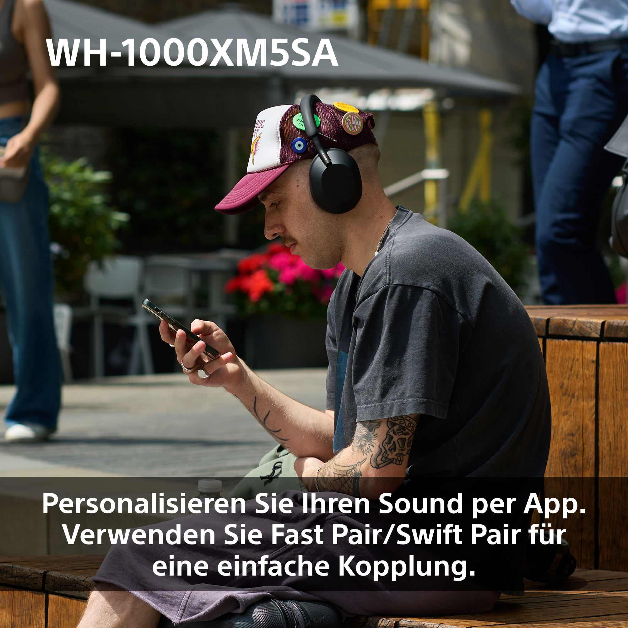 Mann mit Kopfhörern, hält ein Telefon. Er sitzt auf einer Bank. Im Hintergrund sind Menschen, Gebäude und Blumen.