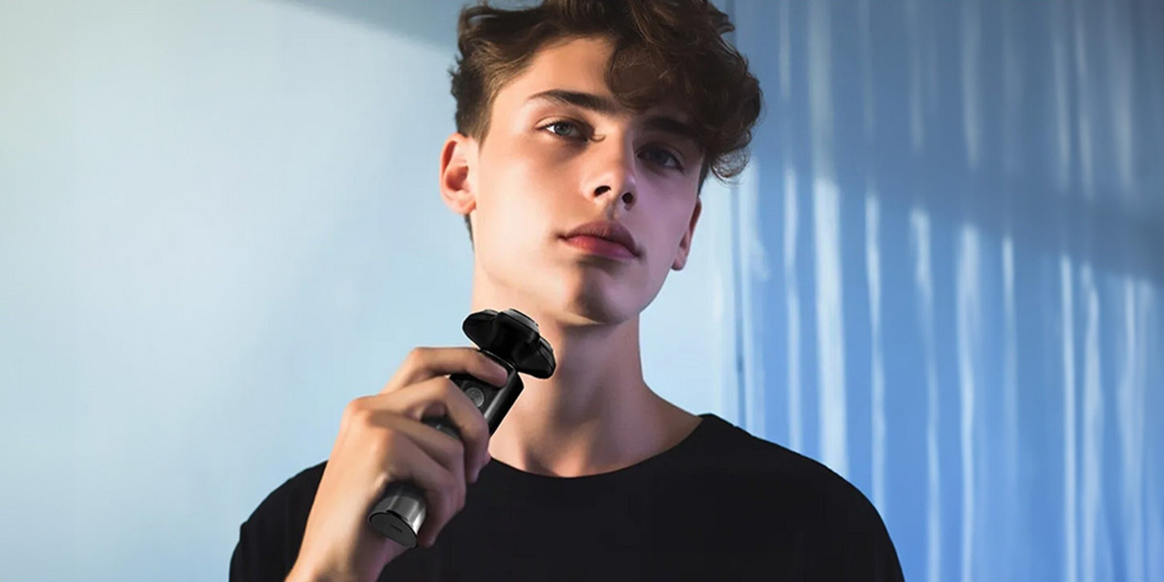 Młody mężczyzna trzyma golarkę elektryczną. Patrzy w kamerę na jasnoniebieskim tle.