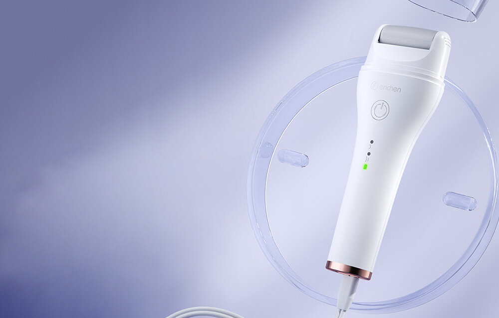 Białe urządzenie do pielęgnacji stóp z szarym wałkiem jest wyświetlane na miękkim fioletowym tle. Spoczywa na przezroczystym plastikowym dysku. Urządzenie posiada przycisk zasilania.