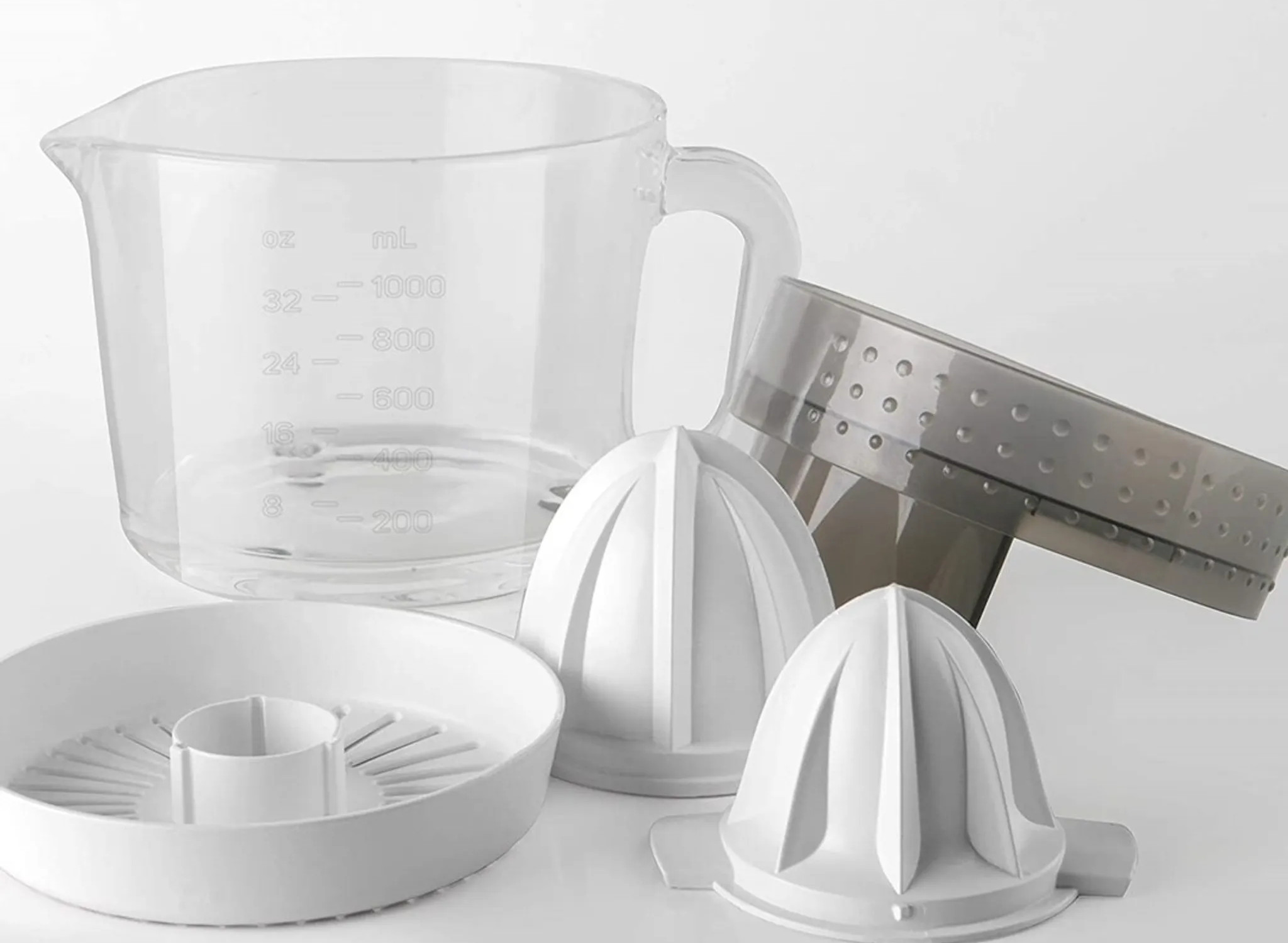 Przezroczysty kubek z miarką i części wyciskarki do cytrusów ułożone na białej powierzchni. Zawiera stożki, sitko i pokrywę.