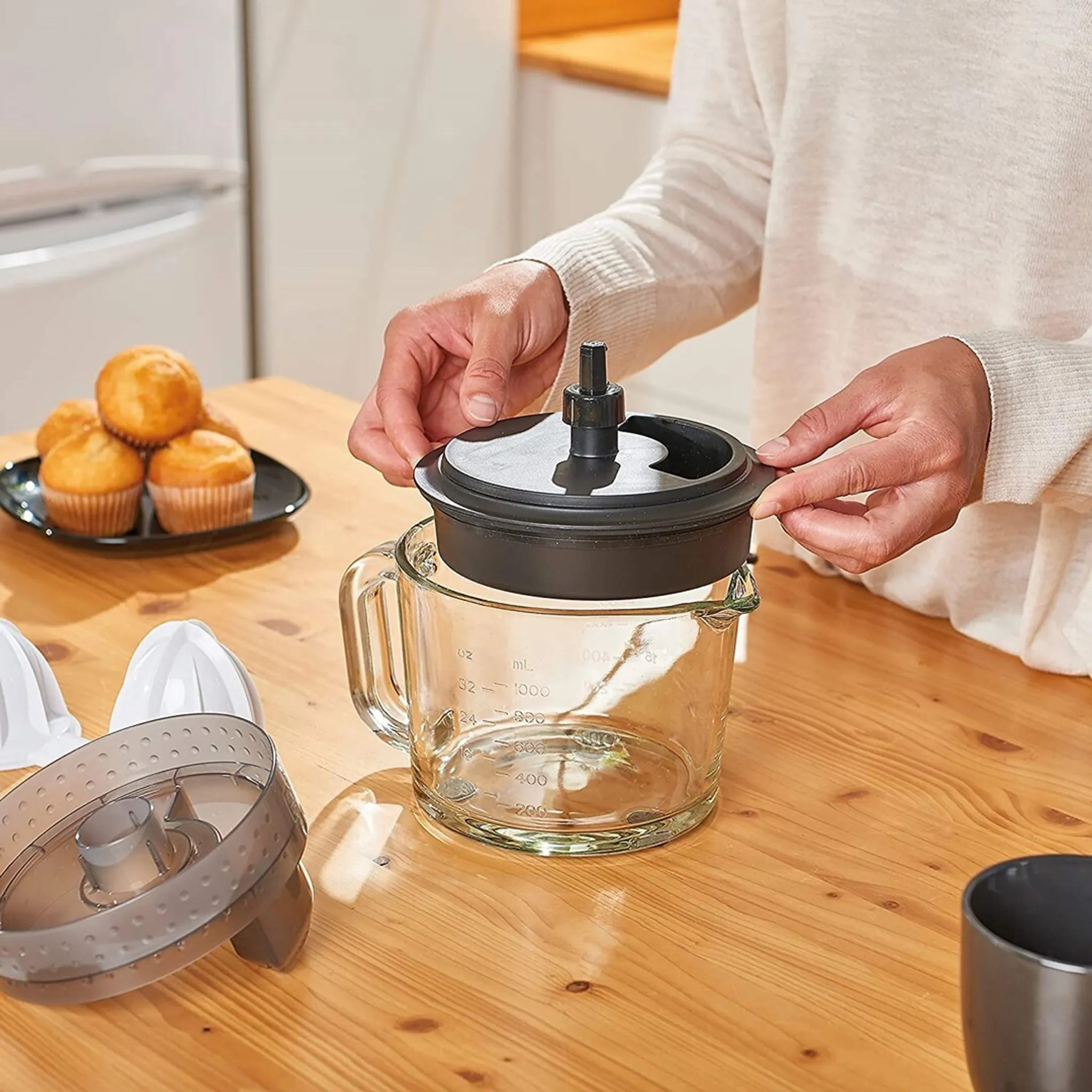 Osoba montuje wyciskarkę. Szklany dzbanek, czarna pokrywa, muffiny i inne części na drewnianym stole.