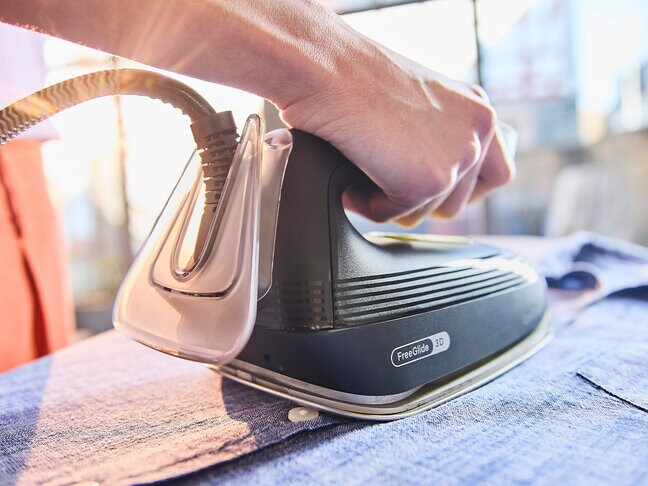 Dłoń osoby prasuje niebieską koszulę czarnym żelazkiem. Żelazko ma przezroczysty zbiornik na wodę. Tło jest rozmyte.