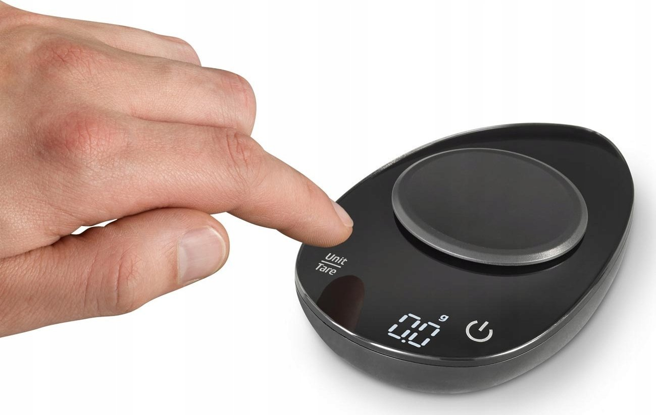 Ręka naciska przycisk na czarnej wadze kuchennej. Waga wyświetla '0,0'. Waga ma owalny kształt.