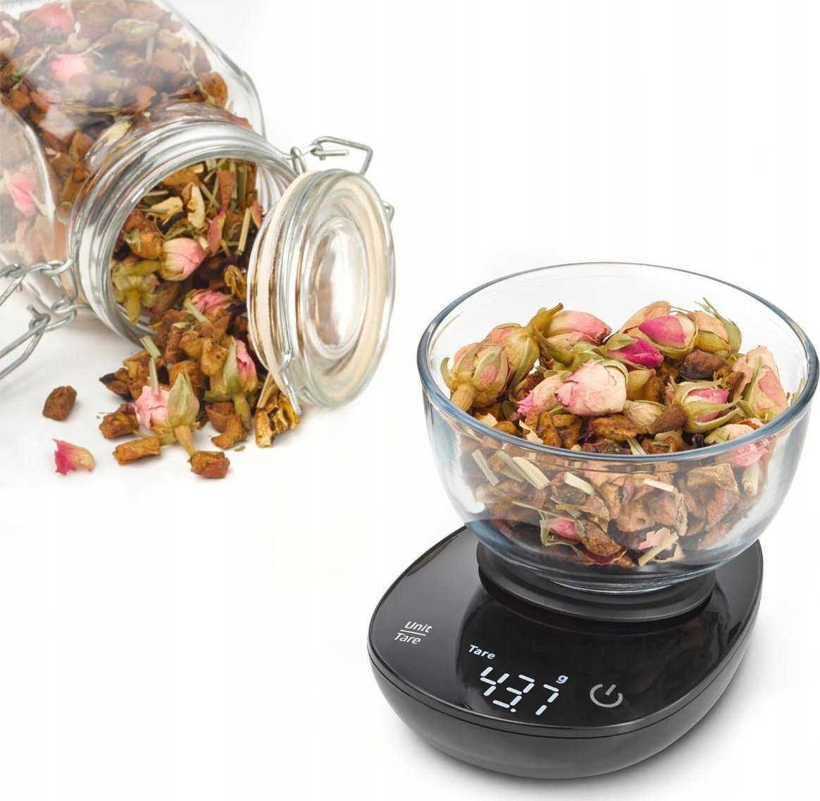 Przezroczysta szklana miska z suszonymi kwiatami na czarnej wadze. Szklany słoik z kwiatami leży obok na białej powierzchni.