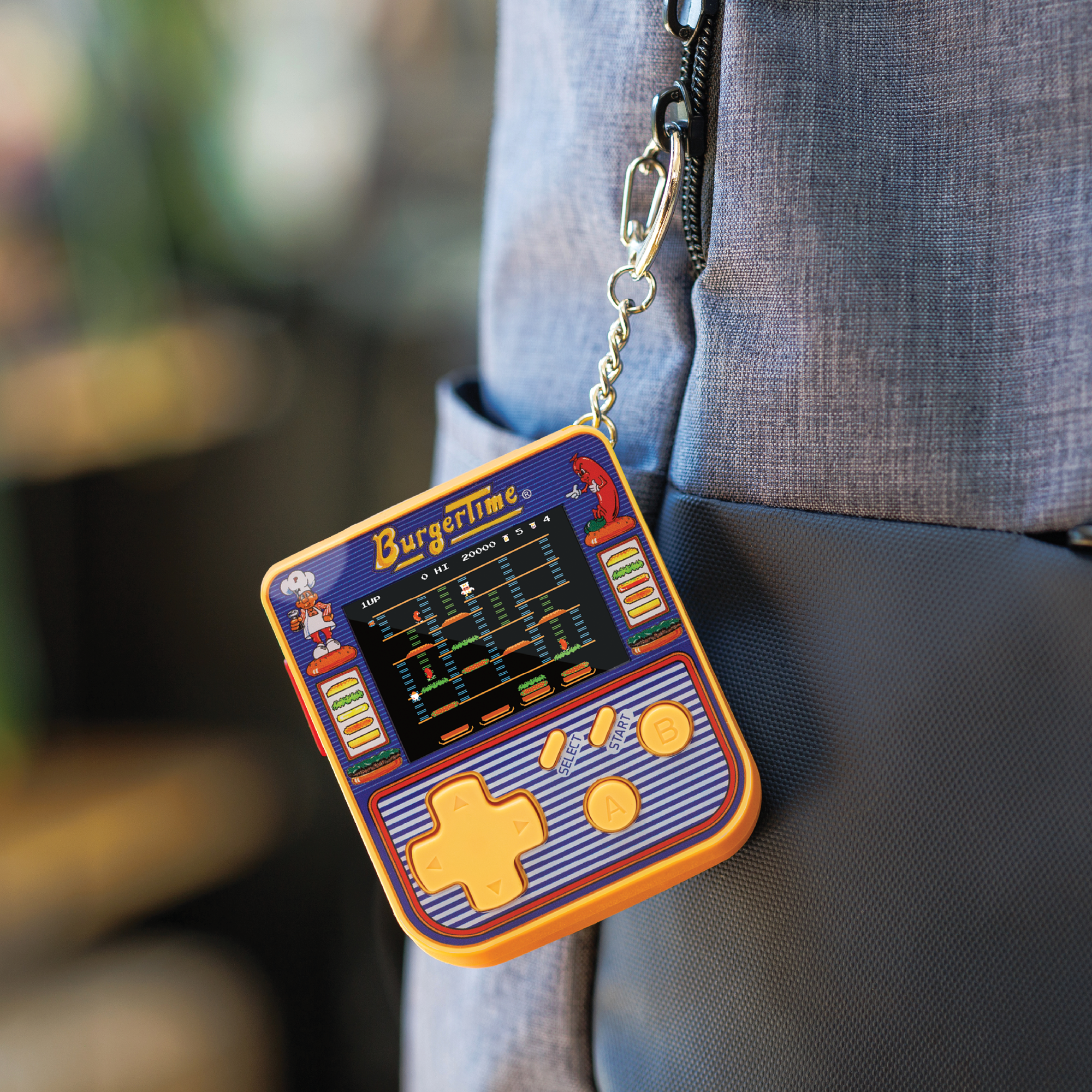 Nahaufnahme eines BurgerTime-Handheld-Spiels, das an einem Rucksack befestigt ist. Das Spiel ist blau und orange und zeigt das Gameplay.