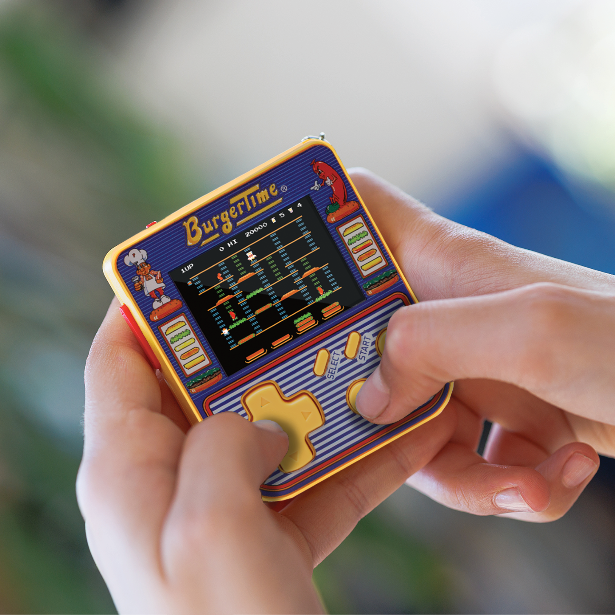 Eine Person hält ein BurgerTime-Handheld-Spiel. Der Bildschirm zeigt das Spiel. Das Gerät ist gelb, blau und rot.