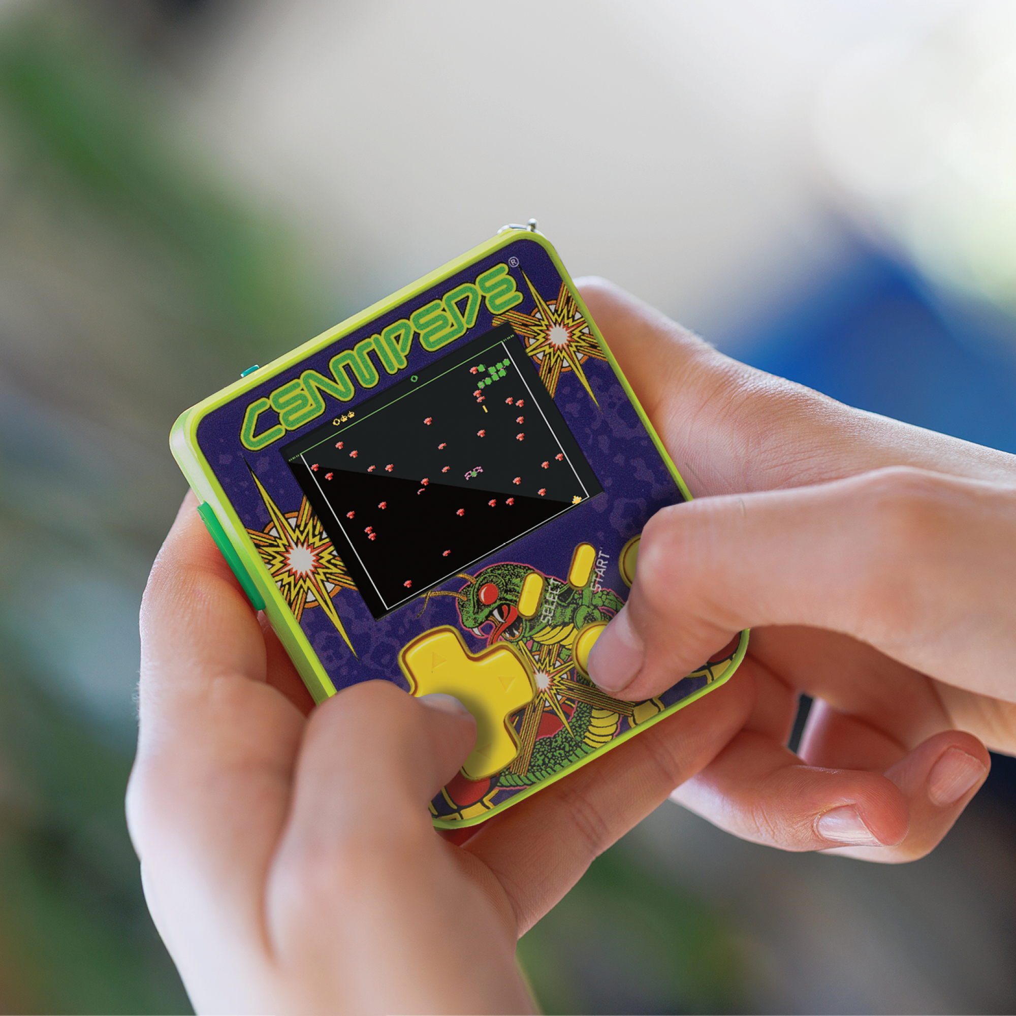 Hände halten ein Centipede-Handheld-Spiel. Der Bildschirm zeigt das Spiel an. Das Gerät ist grün, lila und gelb.